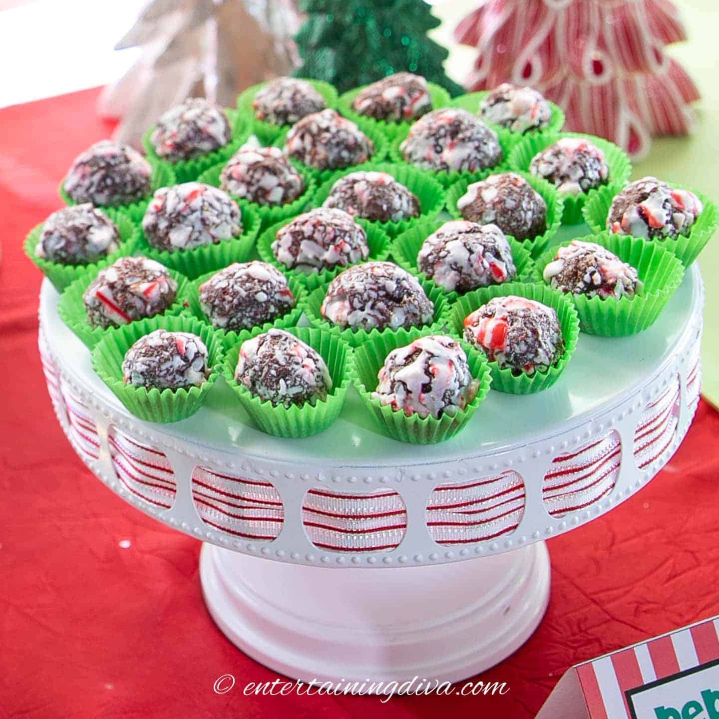 Easy chocolate peppermint fudge balls arranged in green paper cups on a white holiday cake stand, drizzled with white icing and sprinkled with crushed candy canes.