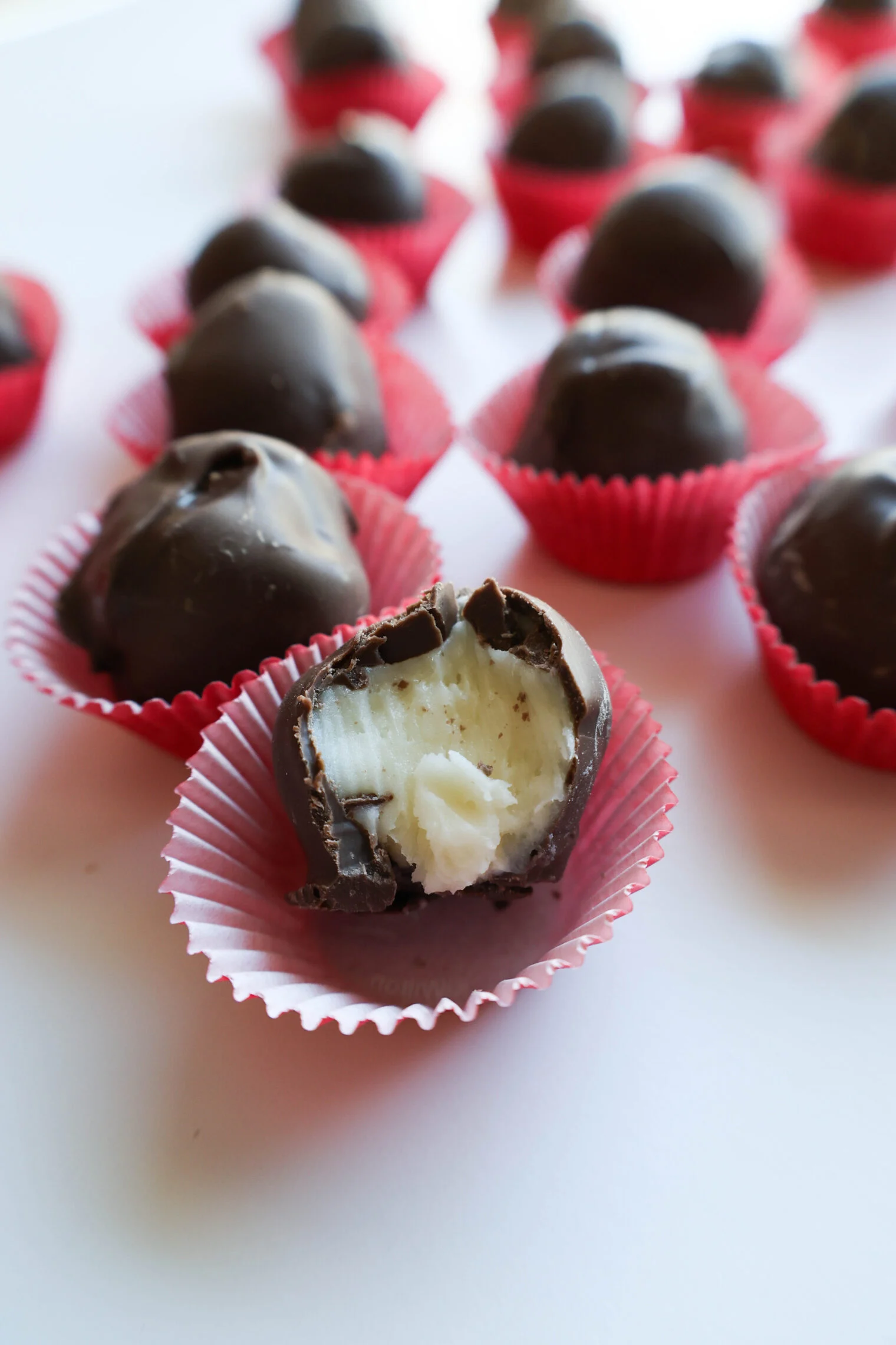 Peppermint buttercream candies with smooth chocolate coating, one with a bite revealing a creamy white peppermint center, displayed in festive red paper cups.