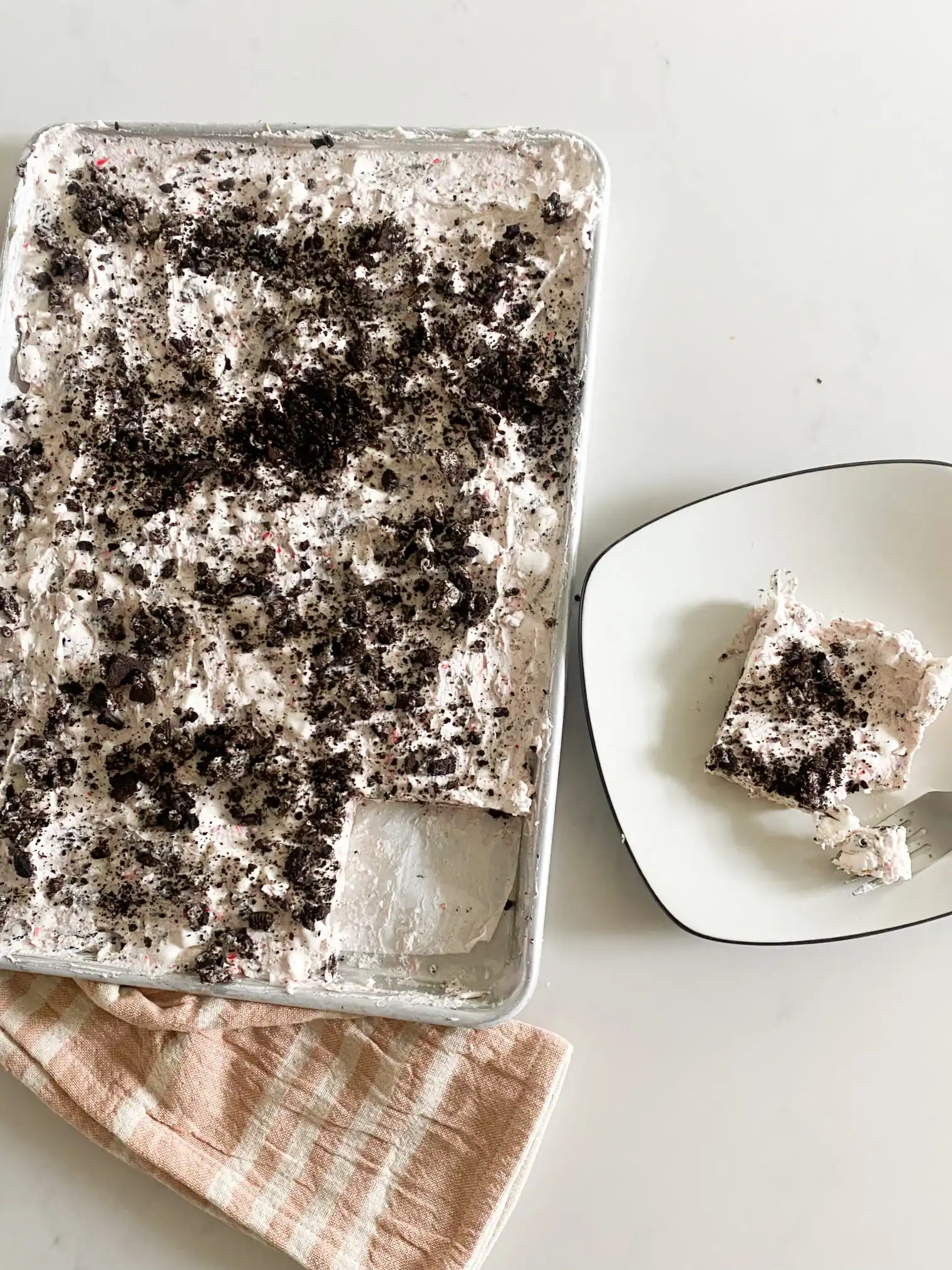 Frozen peppermint dessert topped with whipped cream and crushed Oreos, served in a sheet pan with one square plated beside it on a white background.