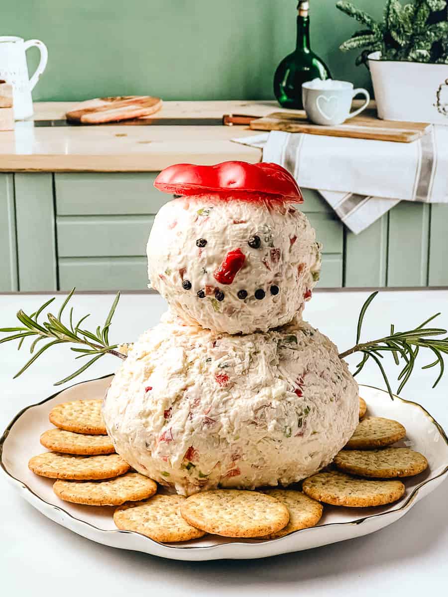 Snowman Cheese Ball made with cream cheese, red peppers, and herbs, shaped into a festive snowman with a red pepper hat, peppercorn eyes, and rosemary arms, surrounded by crackers.