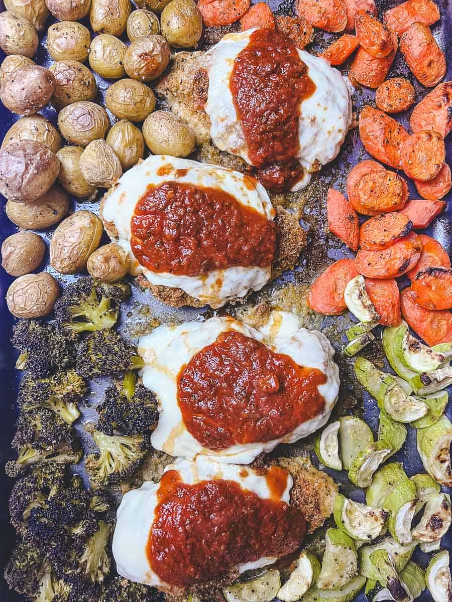 Sheet pan with sheet pan chicken parmesan, parmesan crusted chicken topped with marinara and melted mozzarella, surrounded by roasted vegetables for a one pan meal.