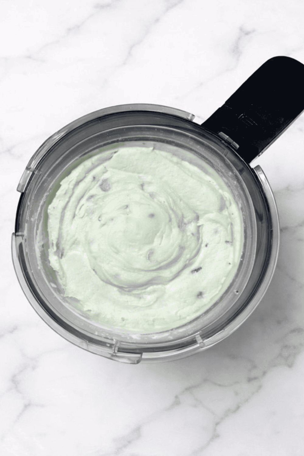 Top-down view of a Ninja Creami pint filled with pale green mint chocolate chip ice cream, set on a clean white marble countertop. The surface is smooth and creamy with evenly blended color and visible chocolate chips.