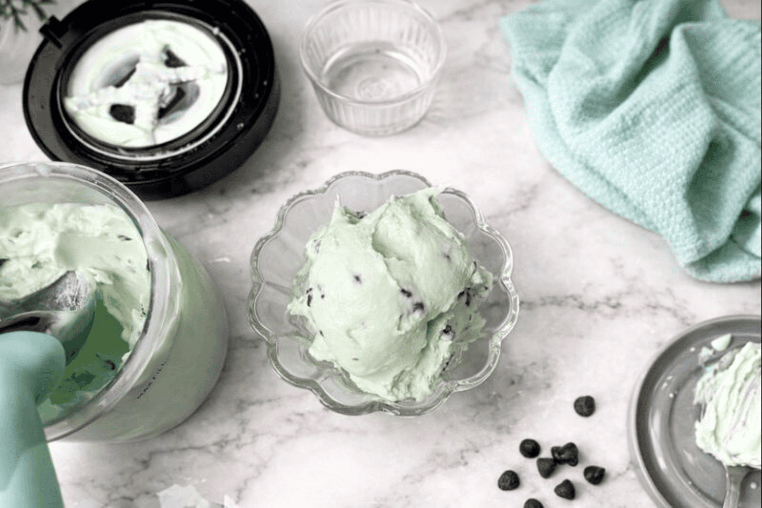 A bowl of creamy mint chocolate chip ice cream sits in a scalloped glass dish on a marble surface, surrounded by a Ninja Creami pint, mint-colored scoop, empty ramekin, and chocolate chips, styled with a folded teal kitchen towel for a cozy, clean aesthetic.