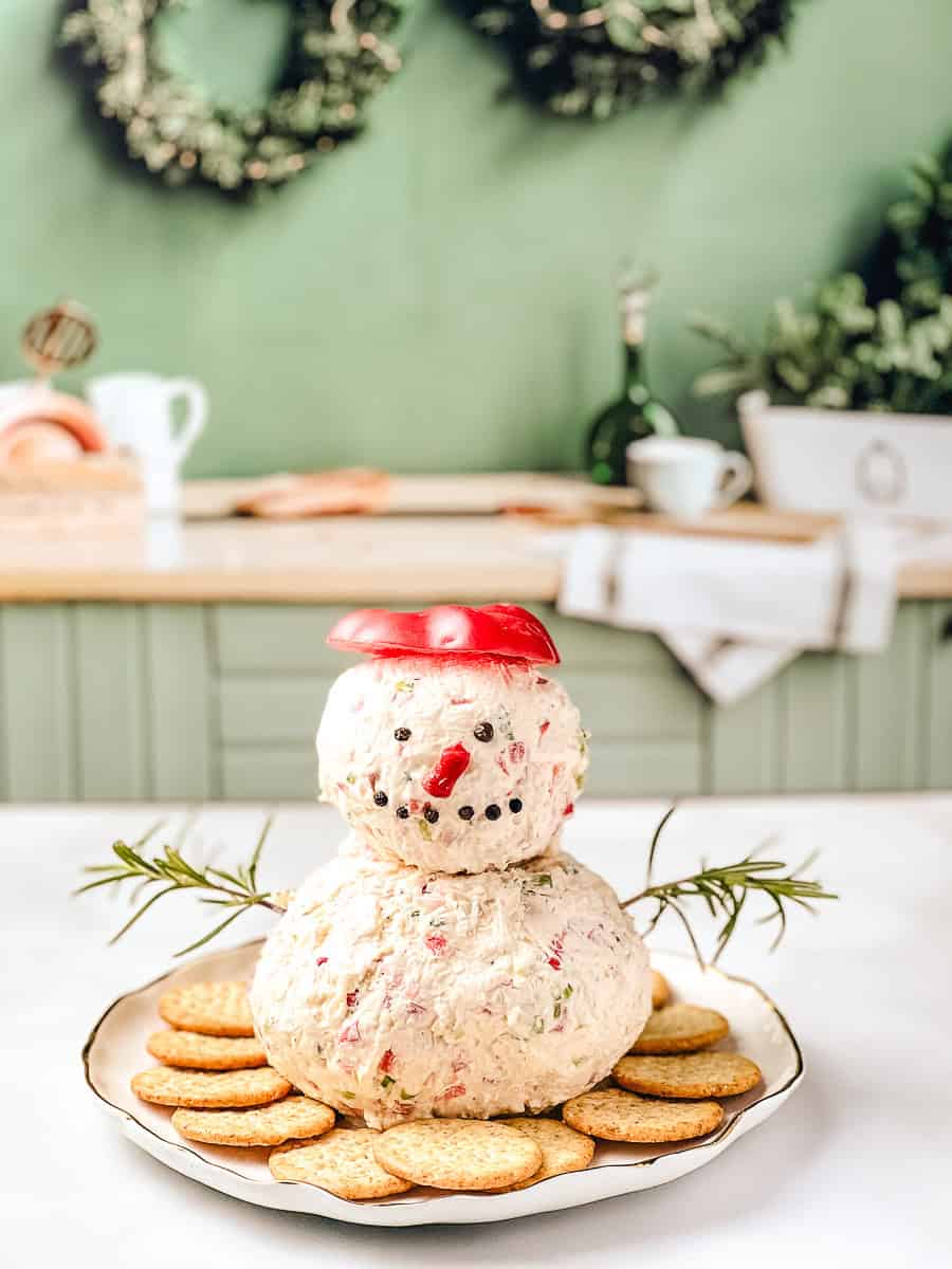 Snowman holiday cheese ball made with cream cheese and white cheddar, decorated with red pepper and chives, served on a platter with crackers as an easy holiday appetizer and festive cheese ball recipe.