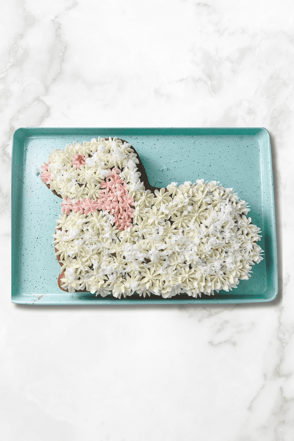 Overhead photo of a lamb-shaped Easter cake decorated with piped white buttercream and coconut on a turquoise tray on a white marble countertop