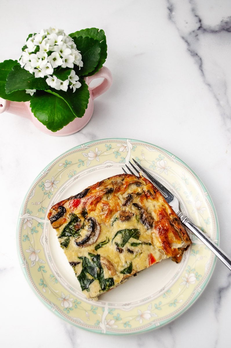 Top-down view of a slice of mushroom spinach egg casserole on a floral plate with a fork, showing browned edges, fluffy baked eggs, sautéed mushrooms, wilted spinach, and small pieces of red pepper on a white marble counter with a pink flower pot in the background