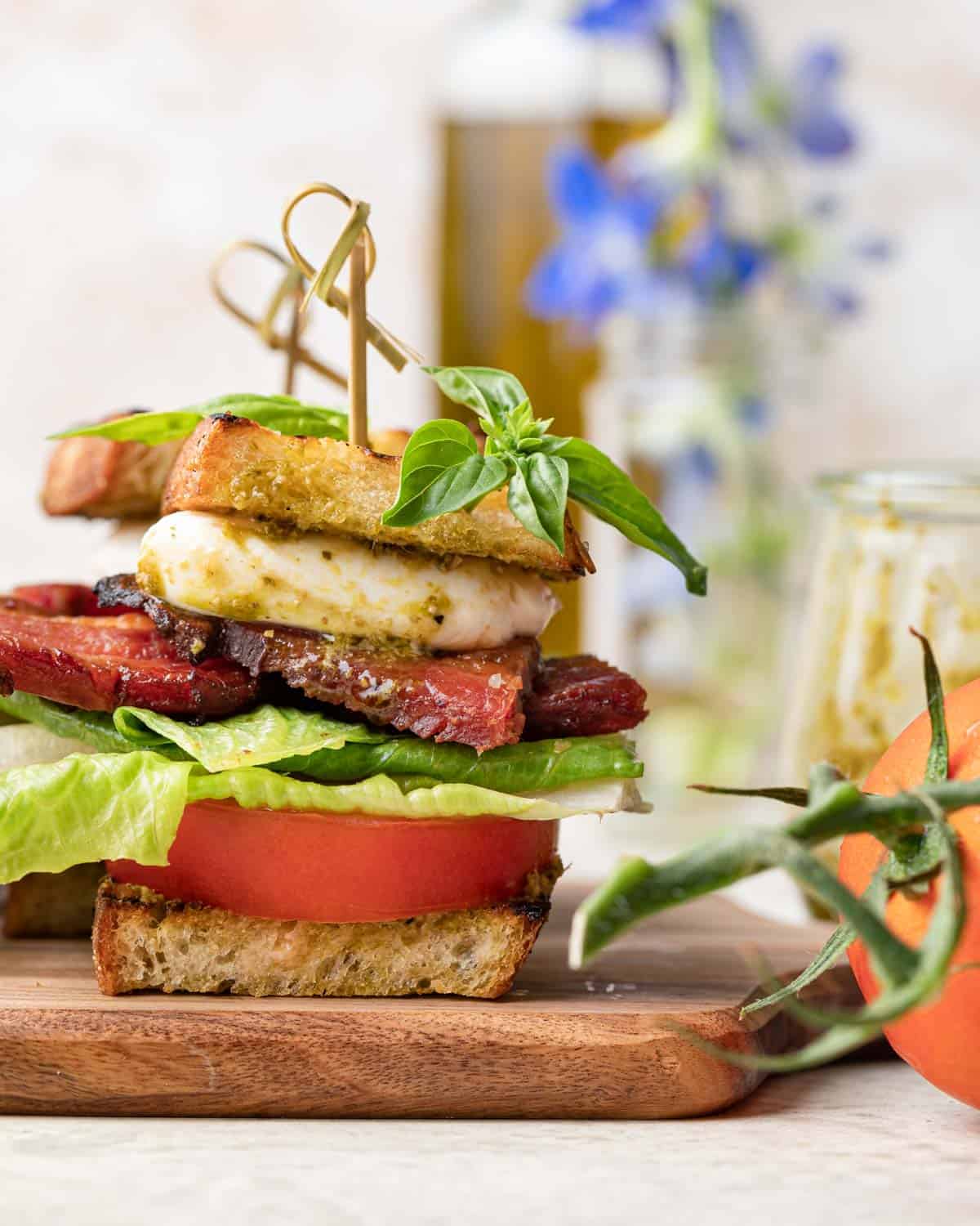 BLT sliders stacked on a wooden cutting board with crispy bacon, fresh tomato slices, crisp lettuce and fresh basil garnish with blue flowers and vine tomatoes in the background, easy cold graduation party slider ideas for a crowd