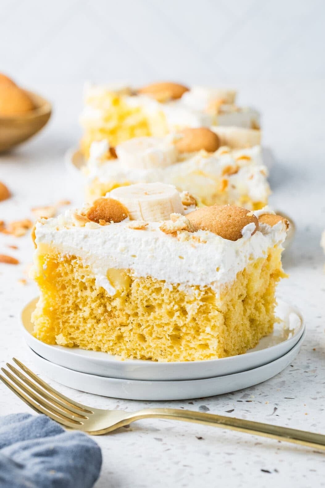 A slice of easy banana pudding poke cake on a stacked white plate with a gold fork, showing a moist yellow cake topped with whipped cream, fresh banana slices, and Nilla wafers, with additional slices visible in the background on a white speckled surface.