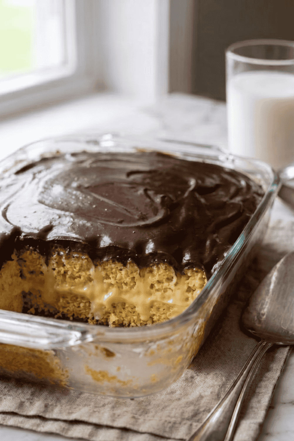 A glass 9x13 baking pan of Boston cream poke cake with one slice removed, showing the creamy vanilla pudding filling inside the golden yellow cake topped with thick glossy chocolate ganache, with a silver spatula and a glass of milk on a linen napkin in the background.