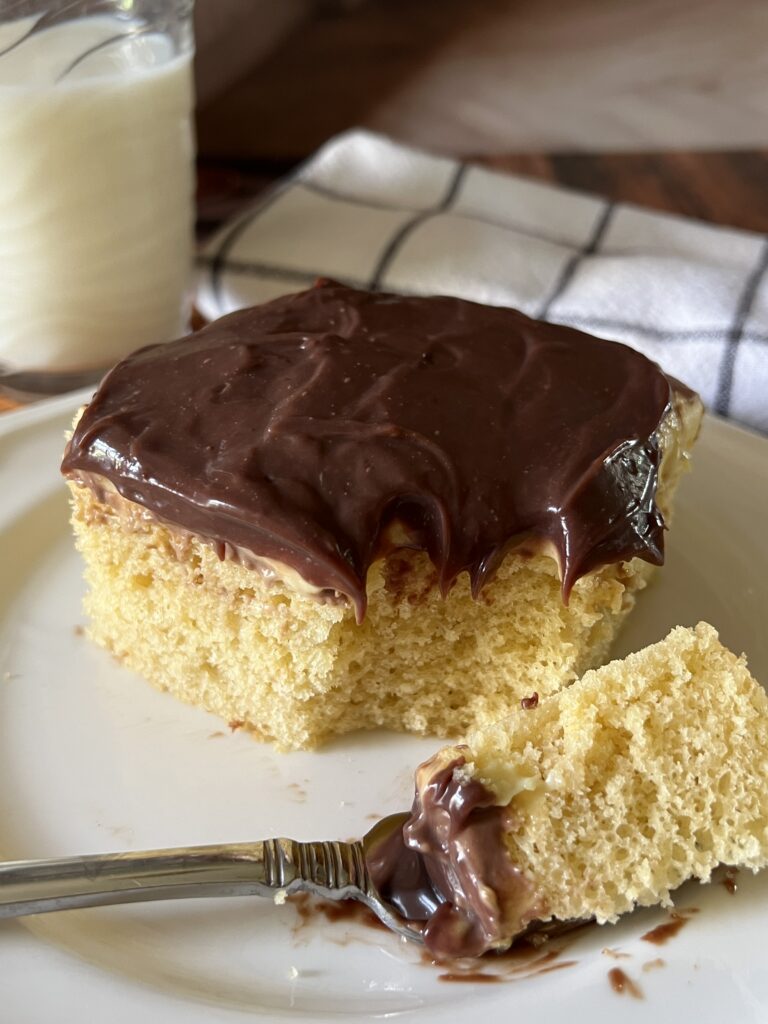 An extreme close-up of a Boston cream poke cake slice on a white plate showing the fluffy yellow cake layers coated in rich glossy chocolate ganache, with a fork loaded with a bite of cake and ganache resting on the plate.