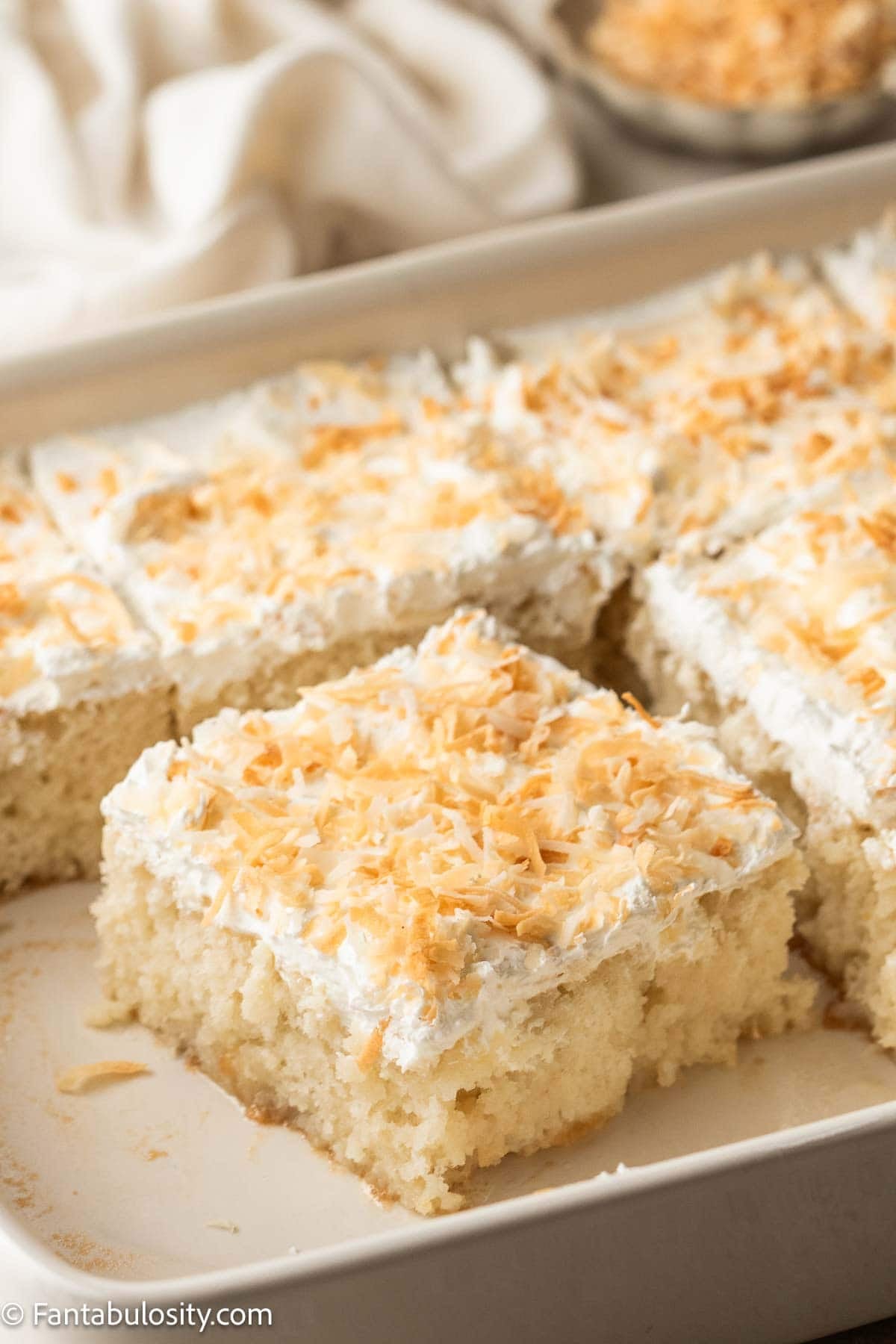 A white baking pan filled with sliced coconut poke cake topped with whipped cream and golden toasted coconut, with a small bowl of toasted coconut and a linen napkin visible in the background.