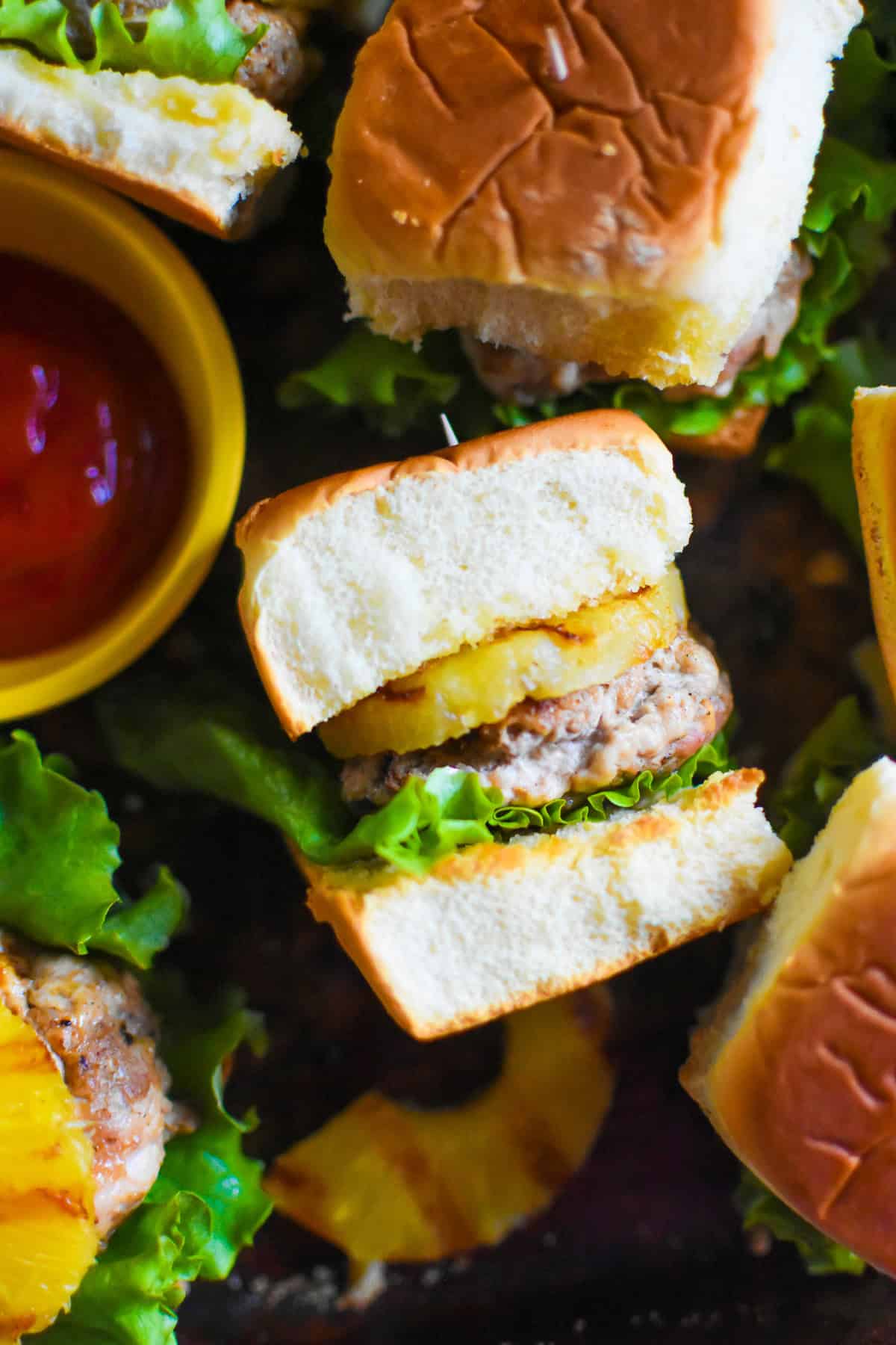 Turkey burger sliders cut in half on King's Hawaiian rolls with grilled pineapple and crisp lettuce on a dark surface with a yellow bowl of ketchup, easy graduation party slider ideas for a crowd