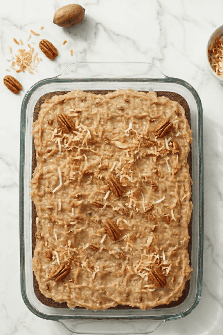 Overhead view of a German Chocolate Poke Cake in a clear glass 9x13 baking dish, topped with coconut pecan frosting, toasted shredded coconut, and whole pecan halves scattered across the top, on a white marble surface with pecans and toasted coconut around the dish.