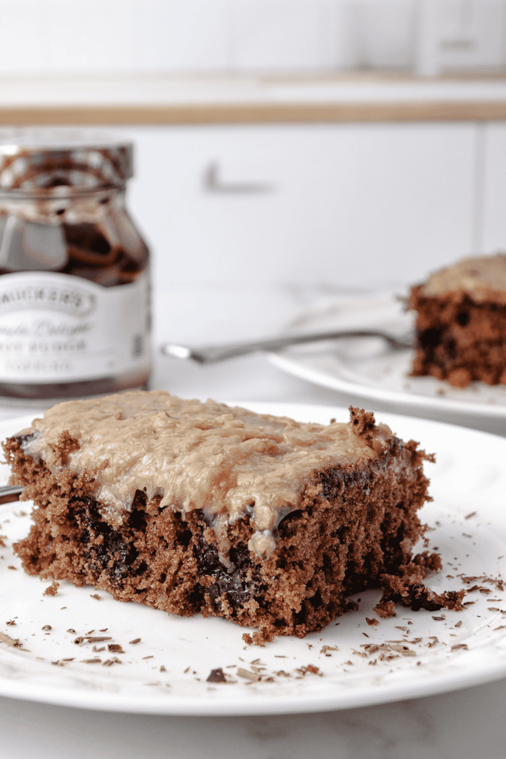 A slice of German Chocolate Poke Cake on a white plate topped with coconut pecan frosting, with a jar of hot fudge and a second slice of cake in the background.