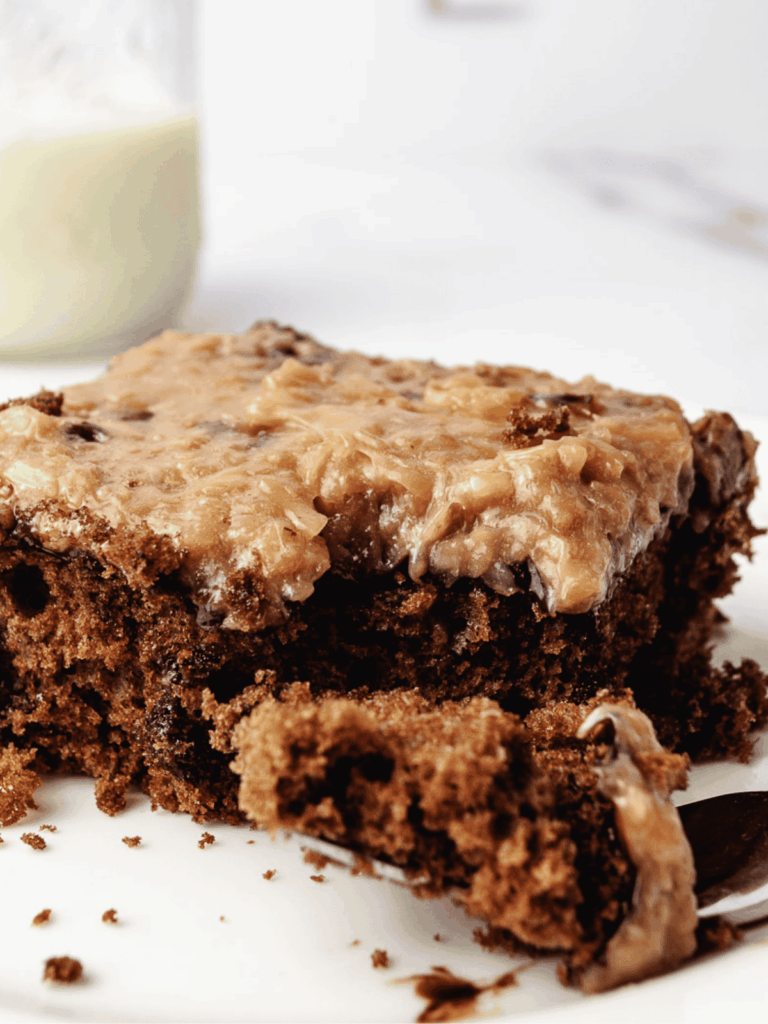 A slice of German Chocolate Poke Cake on a white plate with a fork, topped with coconut pecan frosting, with a glass jar of milk in the background.