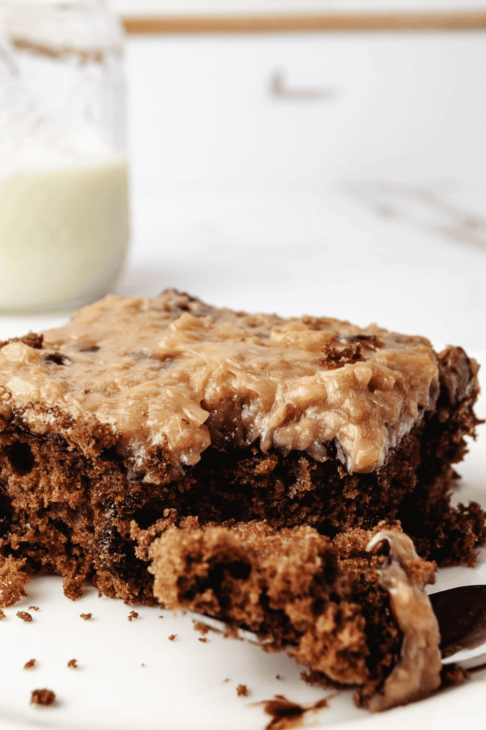 A slice of German Chocolate Poke Cake on a white plate with a fork, topped with coconut pecan frosting, with a glass jar of milk in the background.