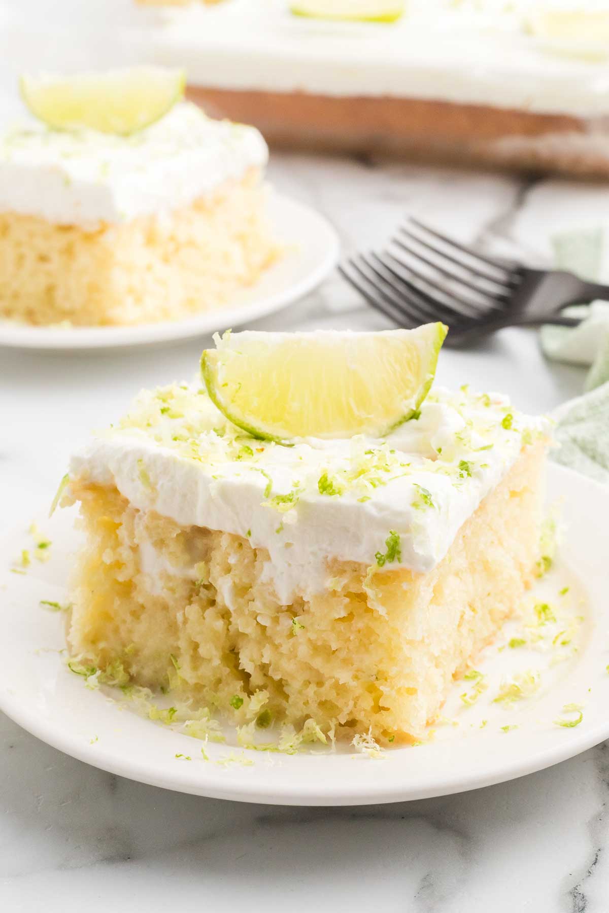 A slice of margarita poke cake on a white plate topped with whipped cream, fresh lime zest, and a lime wedge, with additional slices and a baking pan visible in the background on a marble surface.