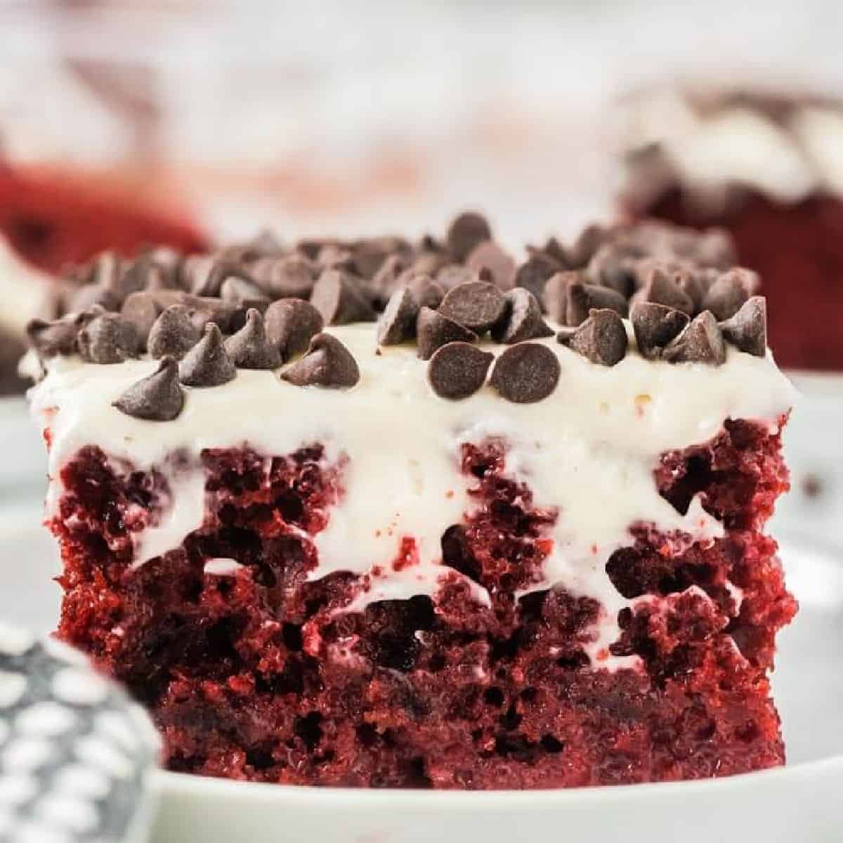 A close-up slice of red velvet poke cake on a white plate, showing a moist deep red cake filled with a creamy white soak and topped with fluffy whipped cream and mini chocolate chips.