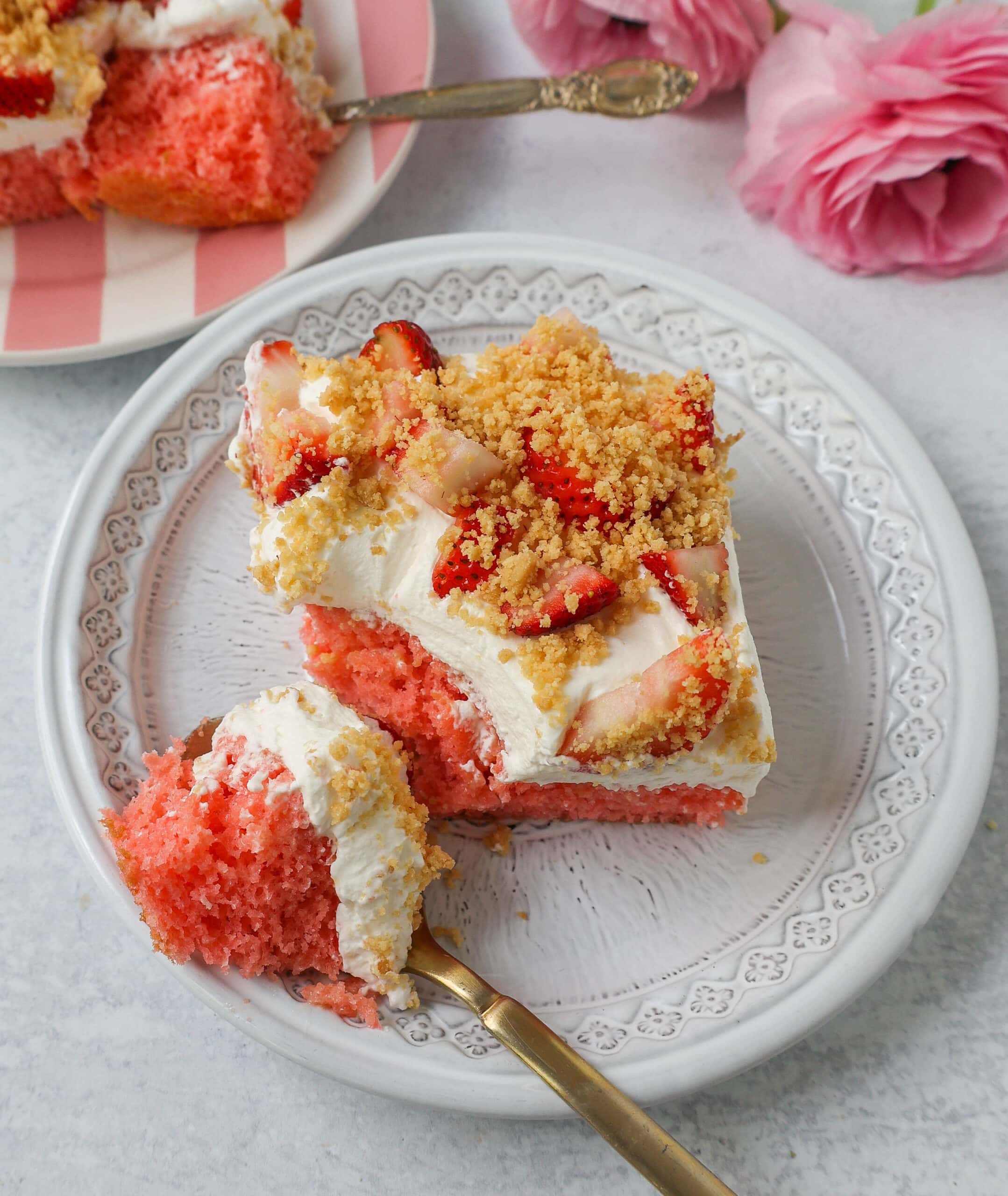 An overhead view of a slice of strawberry cream crunch cake on a decorative white plate with a gold fork, showing a bright pink strawberry poke cake topped with whipped cream, fresh sliced strawberries, and golden graham cracker crumble, with pink roses in the background.
