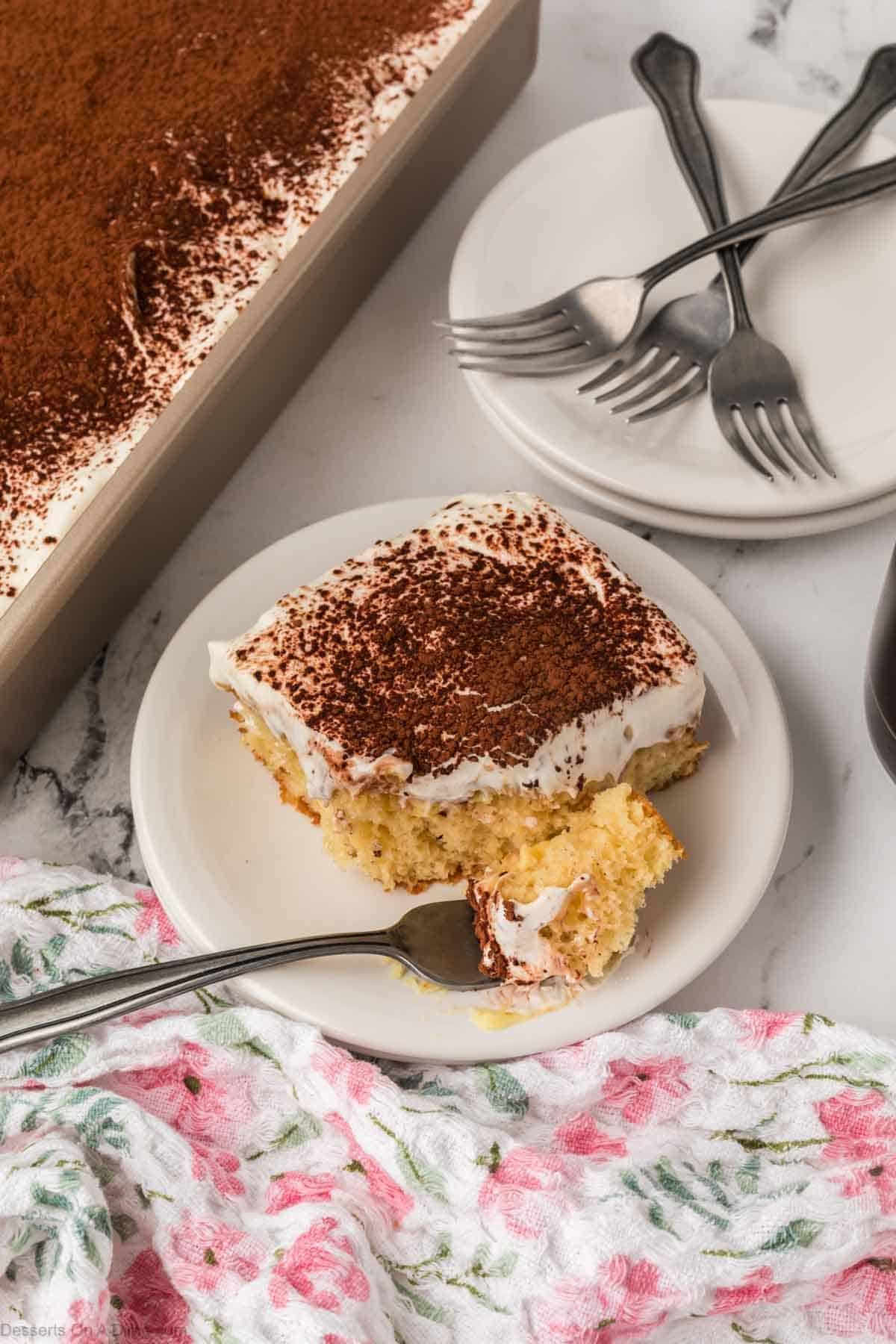 A slice of tiramisu poke cake on a white plate with a fork, showing a golden yellow cake topped with fluffy whipped cream and a dusting of cocoa powder, with the remaining cake in a baking pan on a marble surface in the background.