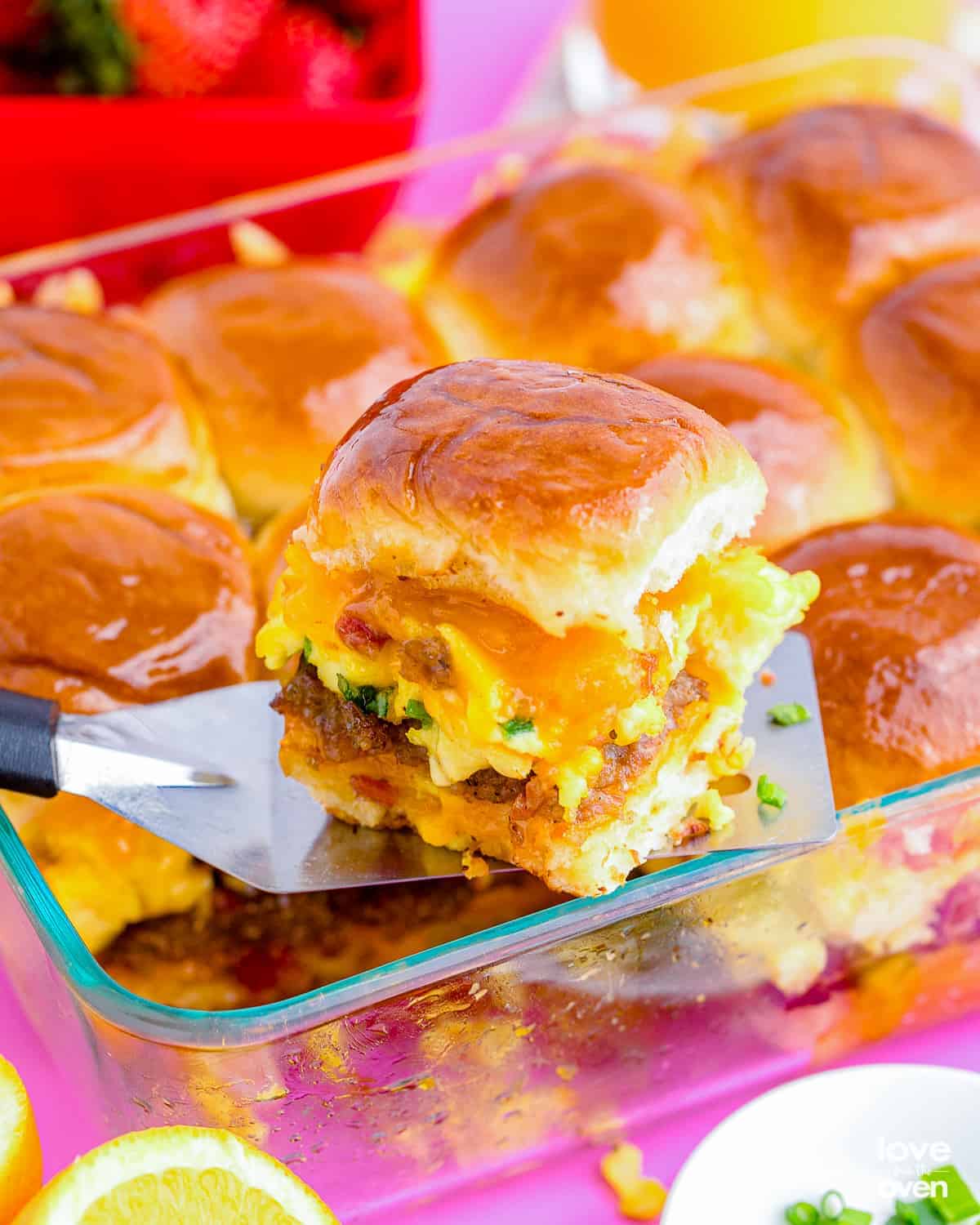 Breakfast sliders with scrambled eggs, sausage, bacon and melted cheddar cheese on soft golden rolls being lifted from a glass baking dish with a spatula, easy make-ahead graduation brunch slider ideas for a crowd