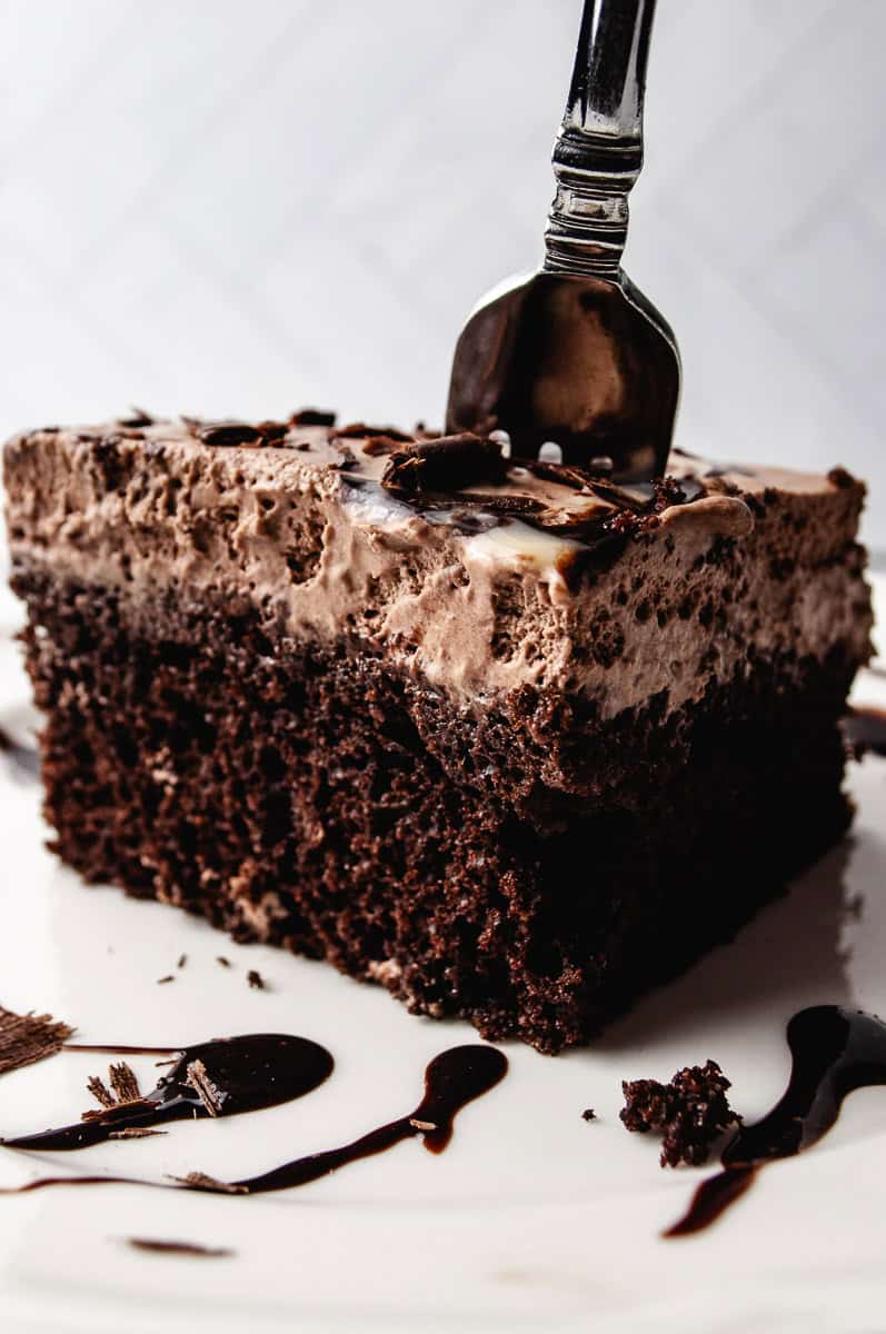 A close up of a tall slice of chocolate poke cake with fluffy chocolate whipped cream frosting, dark chocolate shavings, and a silver fork pressed into the top with chocolate syrup drizzled on a white marble surface