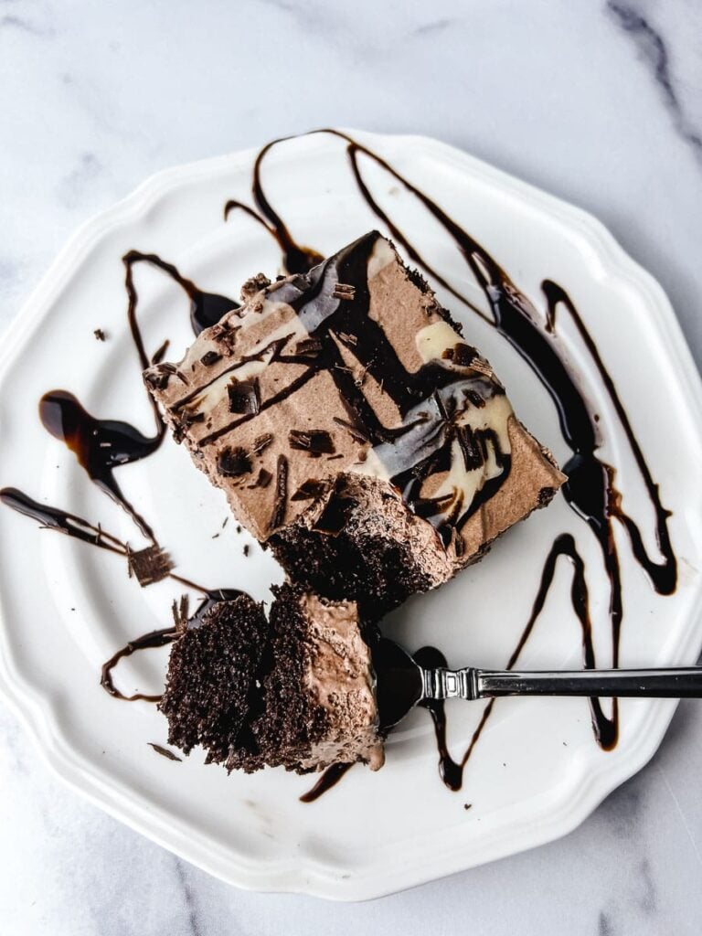 Chocolate poke cake with box cake mix served on a white plate with chocolate syrup drizzle and a fork taking the first bite