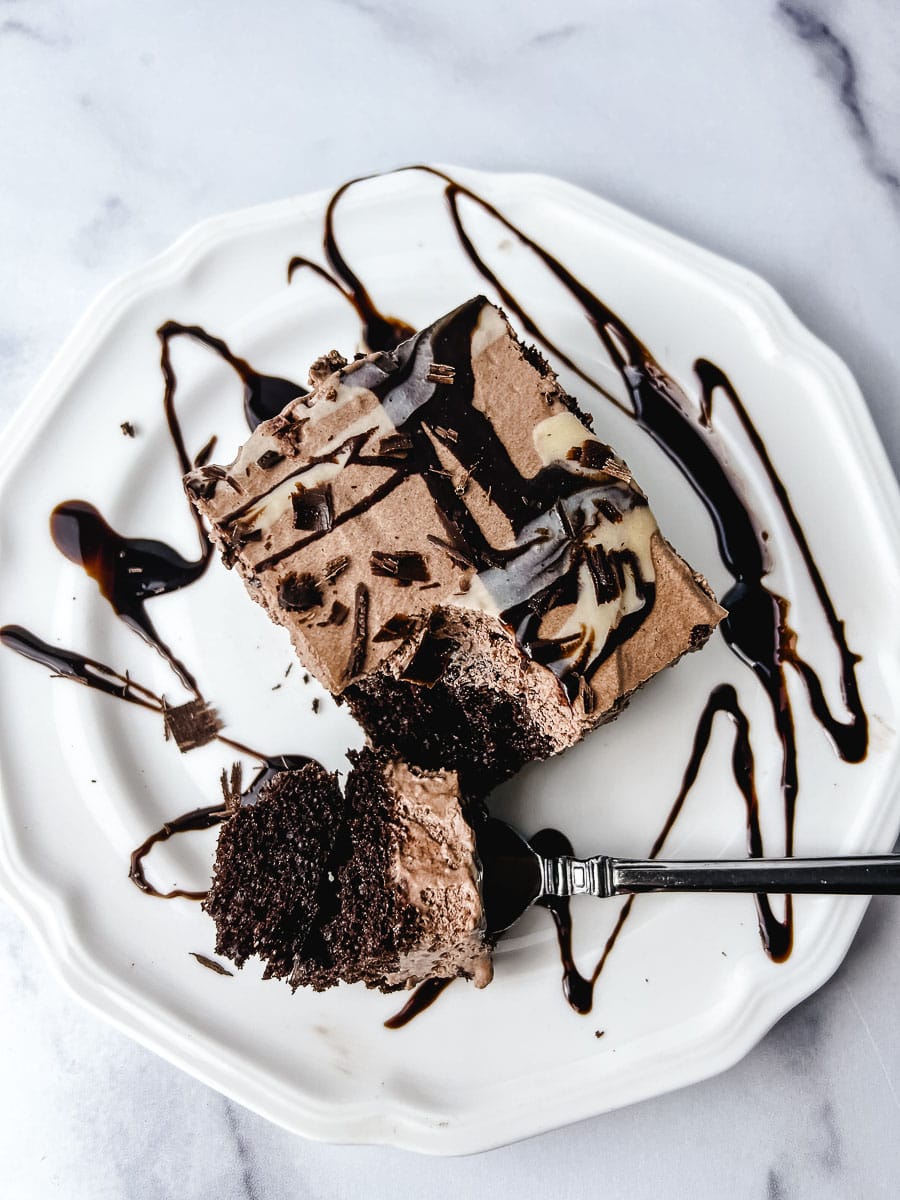 Chocolate poke cake with box cake mix served on a white plate with chocolate syrup drizzle and a fork taking the first bite