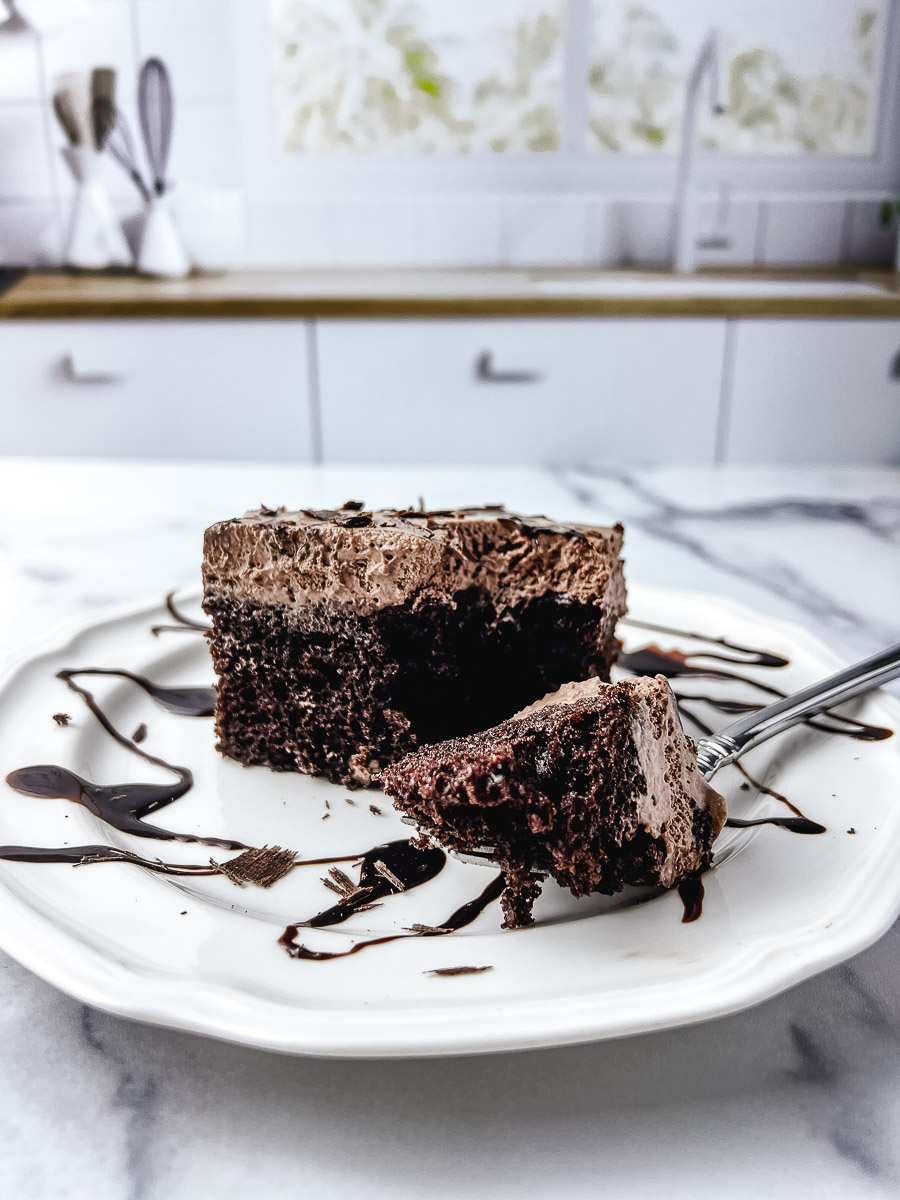 A tall slice of chocolate poke cake topped with a thick layer of fluffy chocolate whipped cream frosting and dark chocolate shavings, served on a white scalloped plate on a marble surface with chocolate syrup drizzled across the plate and a silver fork pulling away a bite, with a bright white kitchen in the background