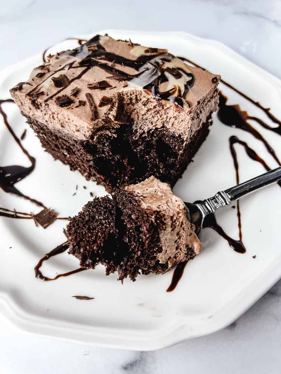 Chocolate poke cake with pudding filling topped with chocolate whipped cream frosting, dark chocolate shavings, and chocolate syrup drizzle on a white plate with a fork taking a bite