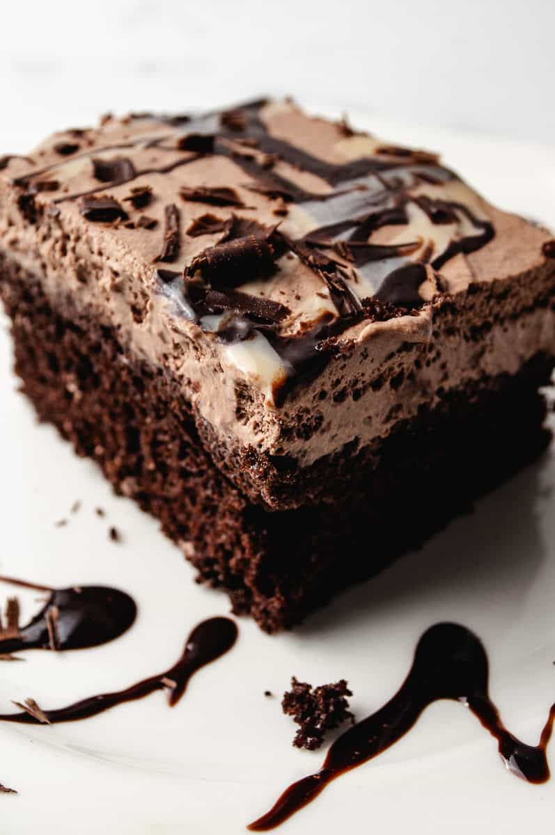 A close up of a slice of chocolate poke cake with pudding topped with fluffy chocolate whipped cream frosting, dark chocolate shavings, and swirls of chocolate and white chocolate syrup on a white plate