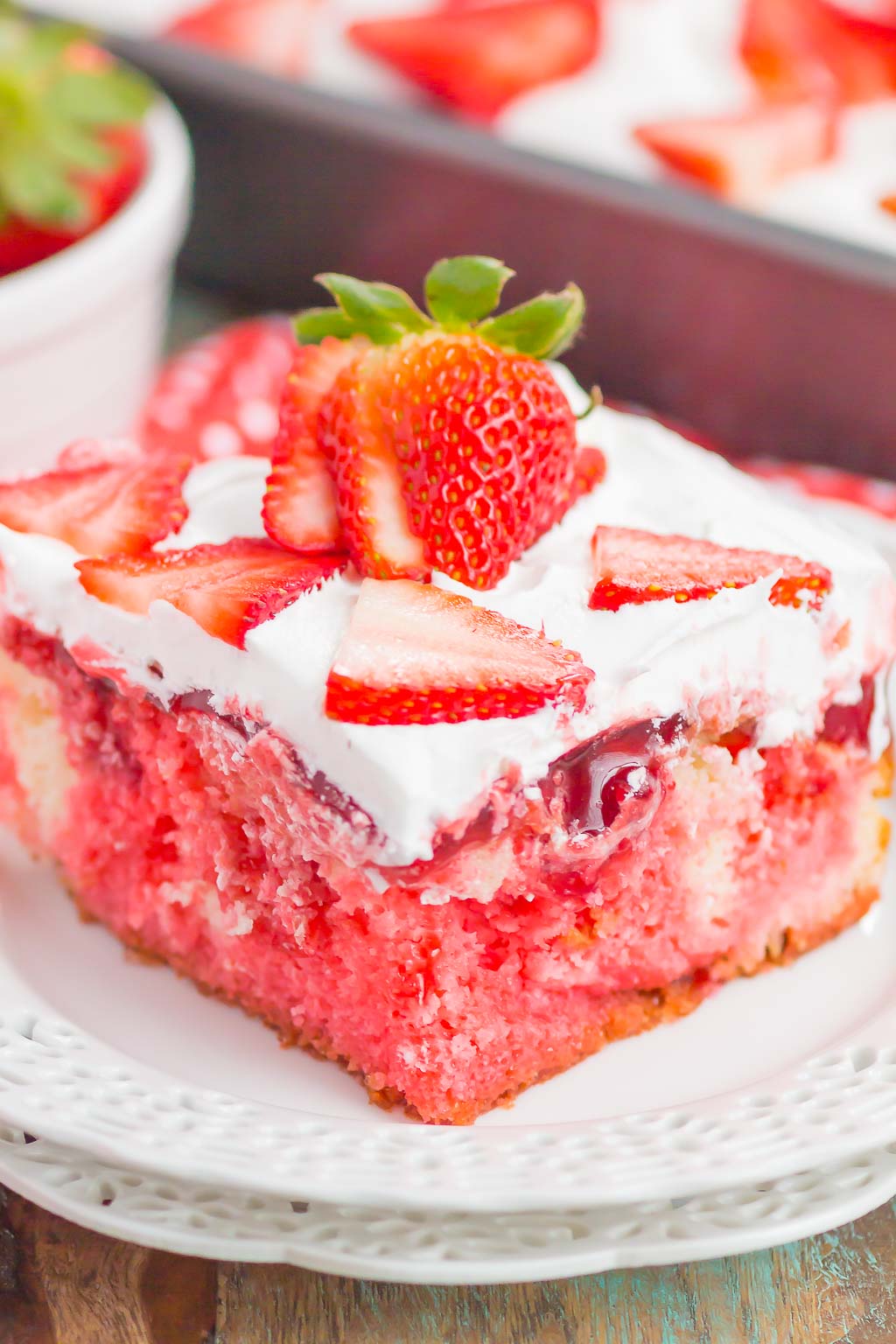 A close-up slice of easy strawberry poke cake on a white lace-edged plate, showing a bright pink jello-soaked cake layered with whipped cream and topped with fresh sliced strawberries and a whole strawberry with the stem on, with the baking pan visible in the background.