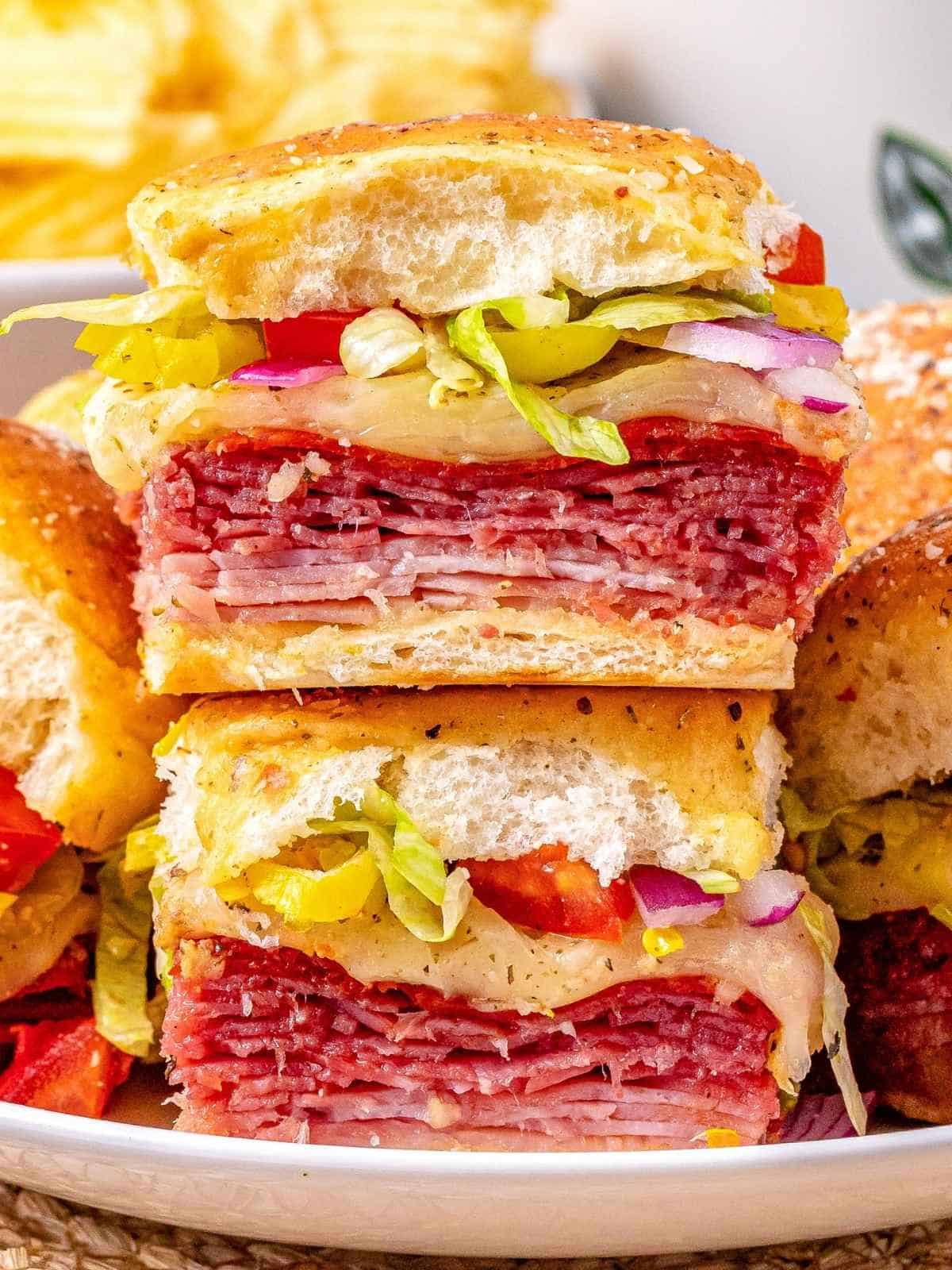 Italian sub sliders stacked and cut in half on a white plate showing layers of salami, ham, pepperoni, melted provolone, shredded lettuce, red onion, red pepper, and pepperoncini on a seasoned roll, easy graduation party slider ideas for a crowd