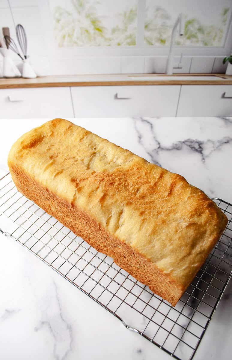 Golden brown homemade bread sourdough white bread loaf cooling on a wire rack on a marble countertop. Easy homemade sandwich bread recipe.