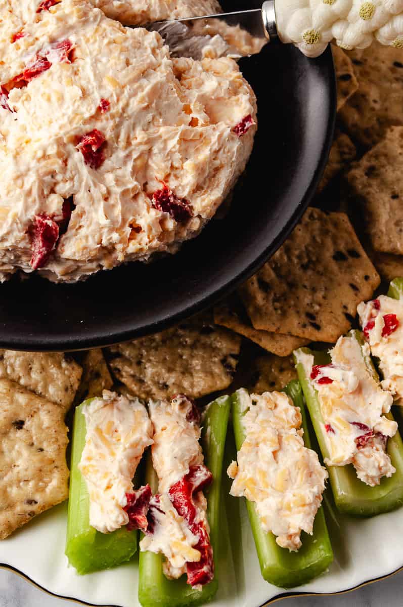 Overhead close-up of homemade Southern pimento cheese being scooped with a silver spreader from a black plate, with fresh celery sticks filled with pimento cheese spread and multigrain crackers arranged on a white scalloped serving platter.
