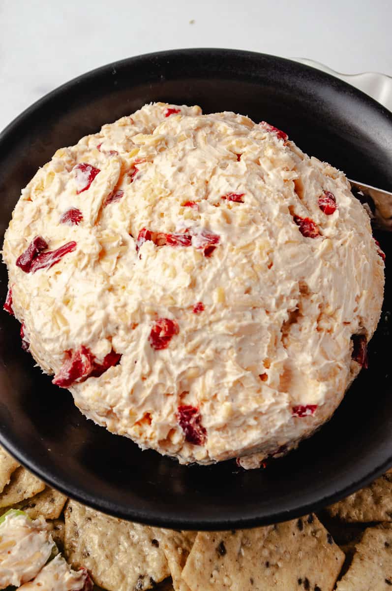 Close-up overhead view of a mound of homemade Southern pimento cheese on a black plate, showing the creamy texture with bright red pimento pieces throughout, with multigrain crackers visible at the bottom edge.