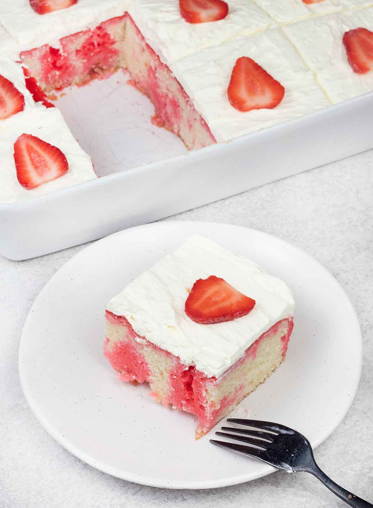 A slice of strawberry poke cake on a white plate with a black fork, showing a white cake filled with bright pink strawberry jello and topped with whipped cream and a fresh sliced strawberry, with the full baking pan visible in the background.