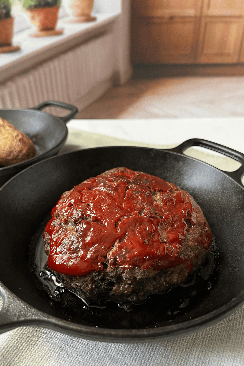 An old fashioned meatloaf with a shiny ketchup topping baked in a black cast iron skillet, photographed on a cream linen with warm wood cabinets and herb pots on a windowsill behind it.