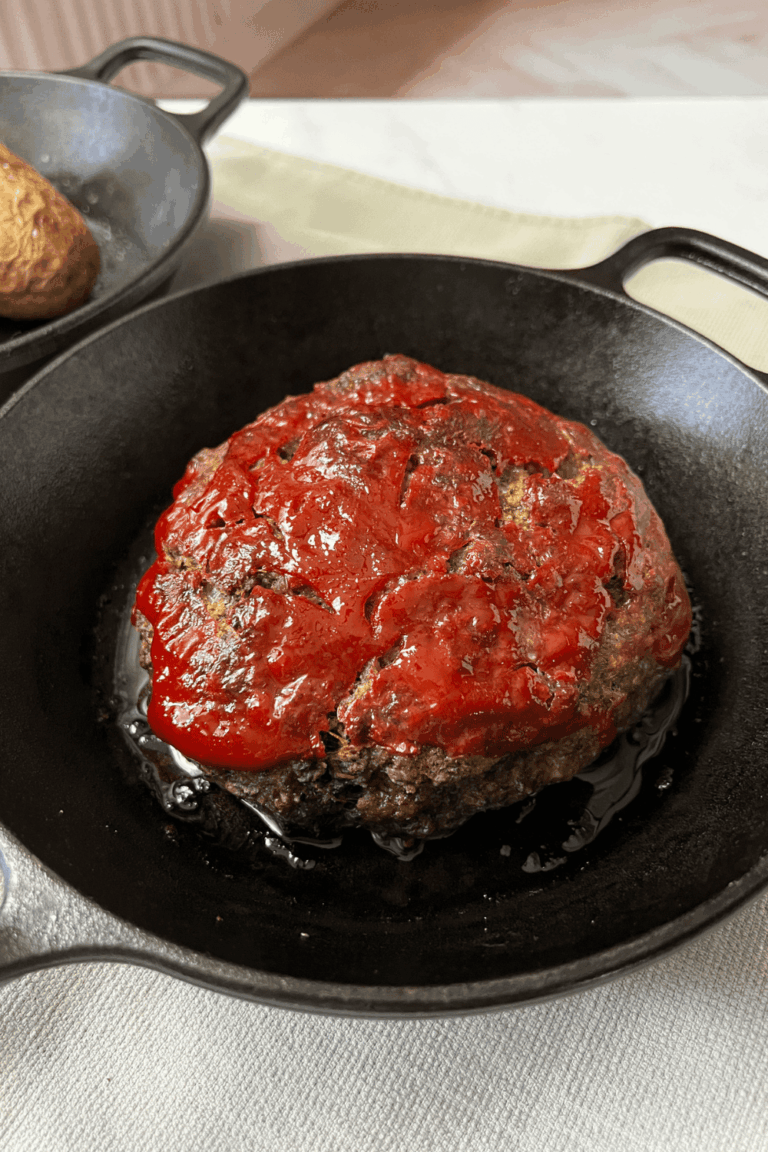 An old fashioned meatloaf with a shiny ketchup topping baked in a black cast iron skillet, photographed on a cream linen