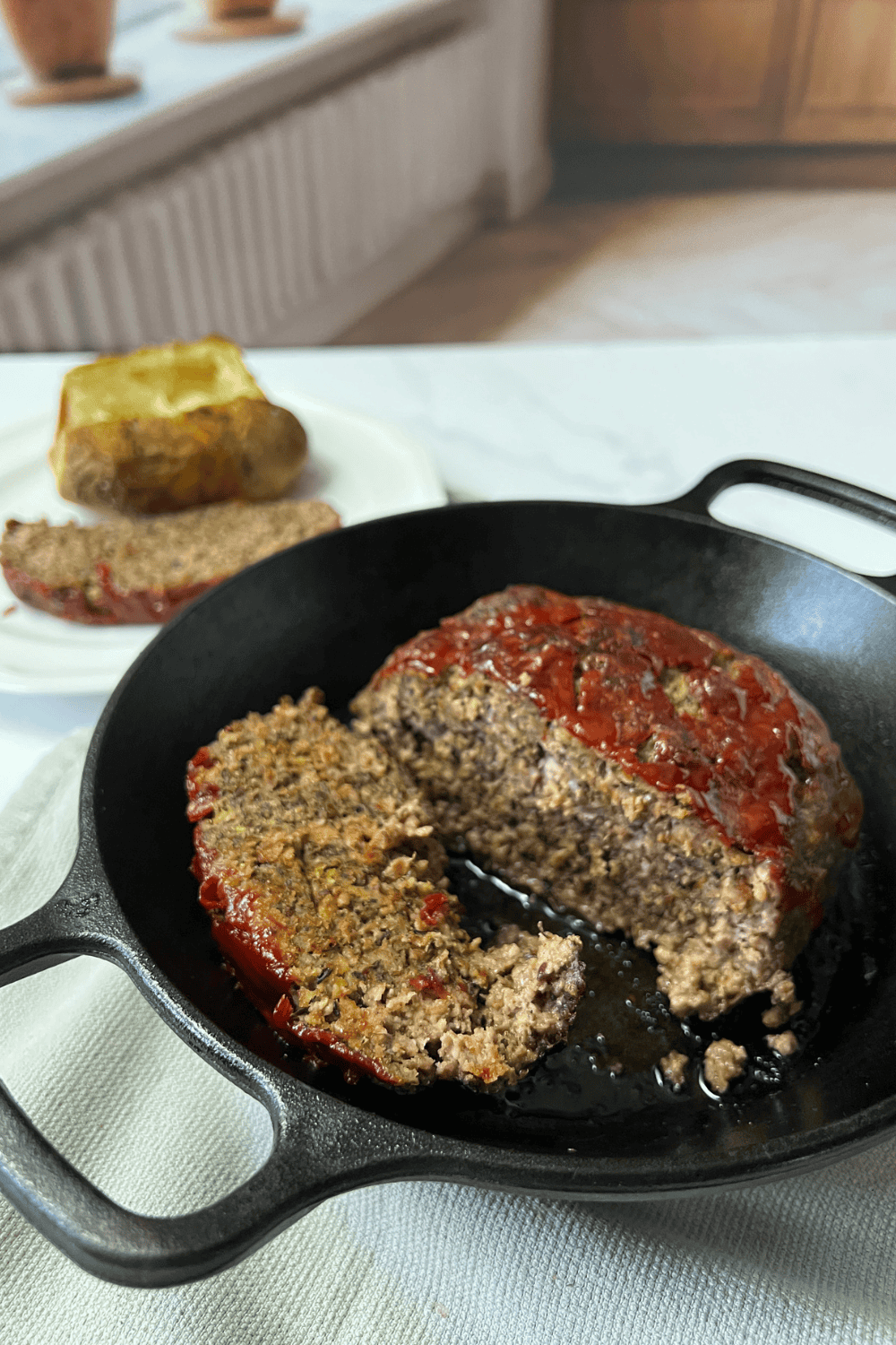 A simple cast iron skillet meatloaf cut open to show the moist, tender inside, with a caramelized ketchup topping, a served slice and baked potato on a white plate in the soft natural light background.