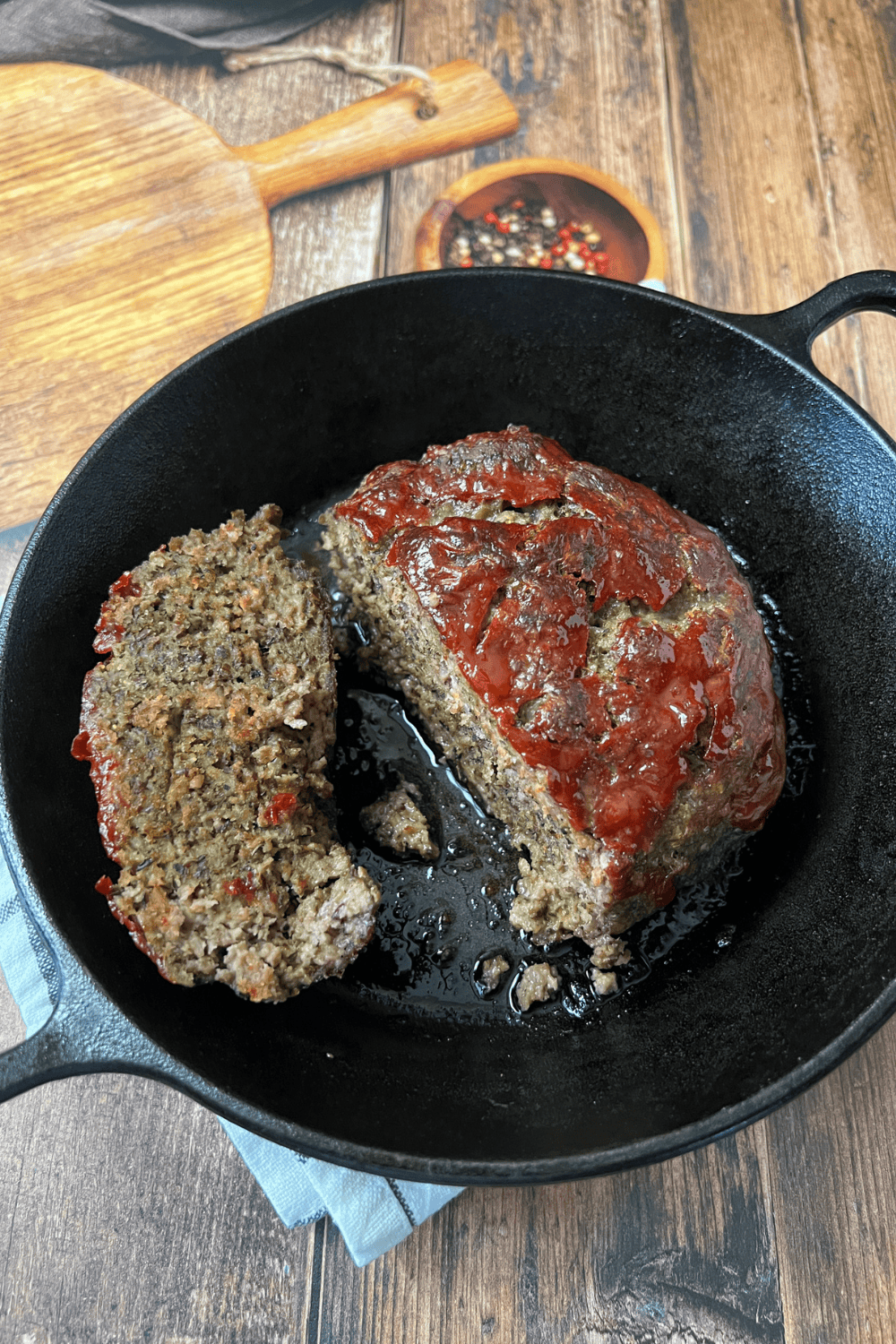 Overhead view of a sliced cast iron skillet meatloaf with a caramelized ketchup glaze, one slice laid flat showing the juicy interior, in a black cast iron skillet on a rustic dark wood surface with a blue linen and a small bowl of peppercorns in the background.