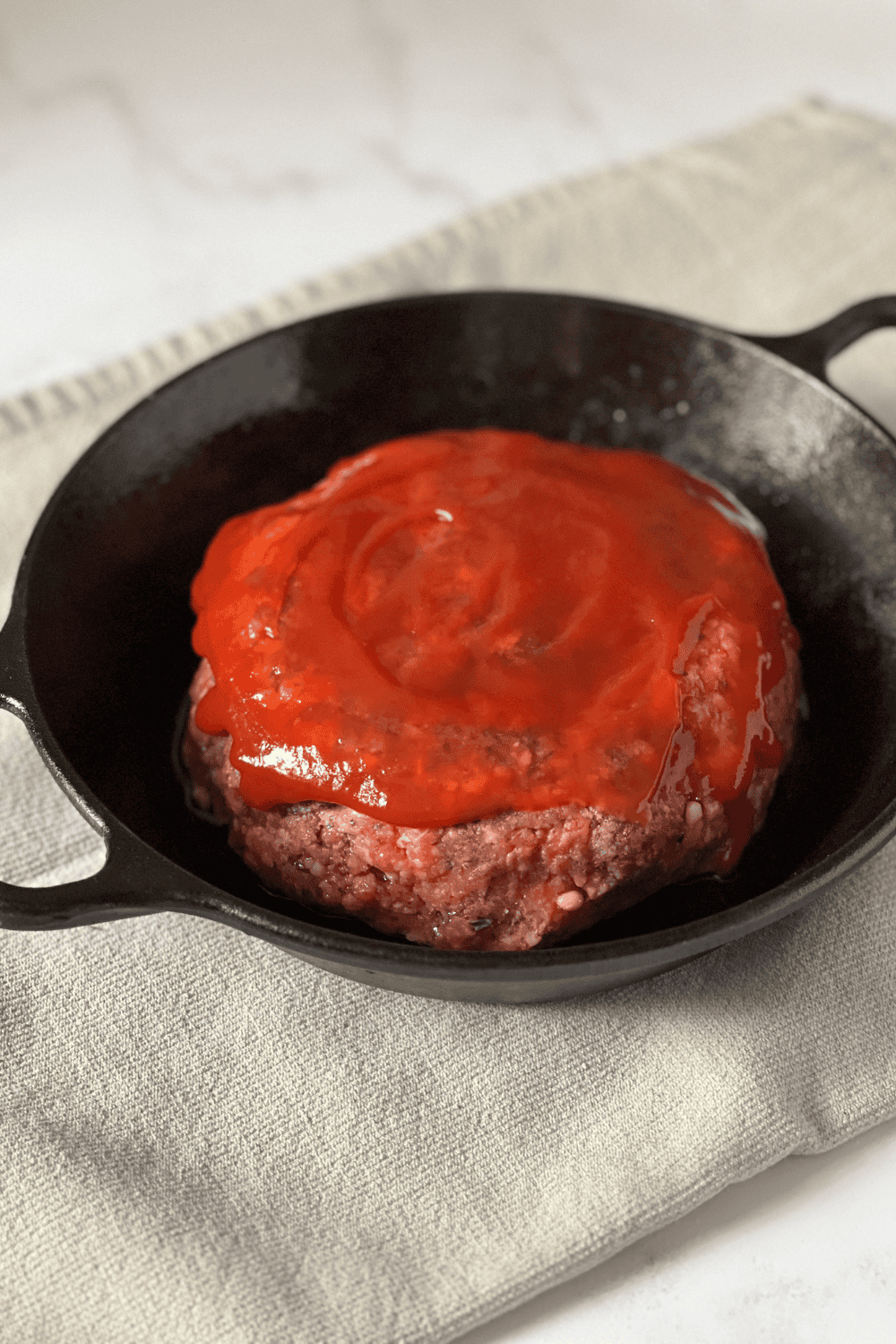 Raw cast iron skillet meatloaf shaped into a round loaf with bright red ketchup swirled on top, sitting in a black cast iron skillet on a cream linen cloth over a white marble surface, ready to bake.