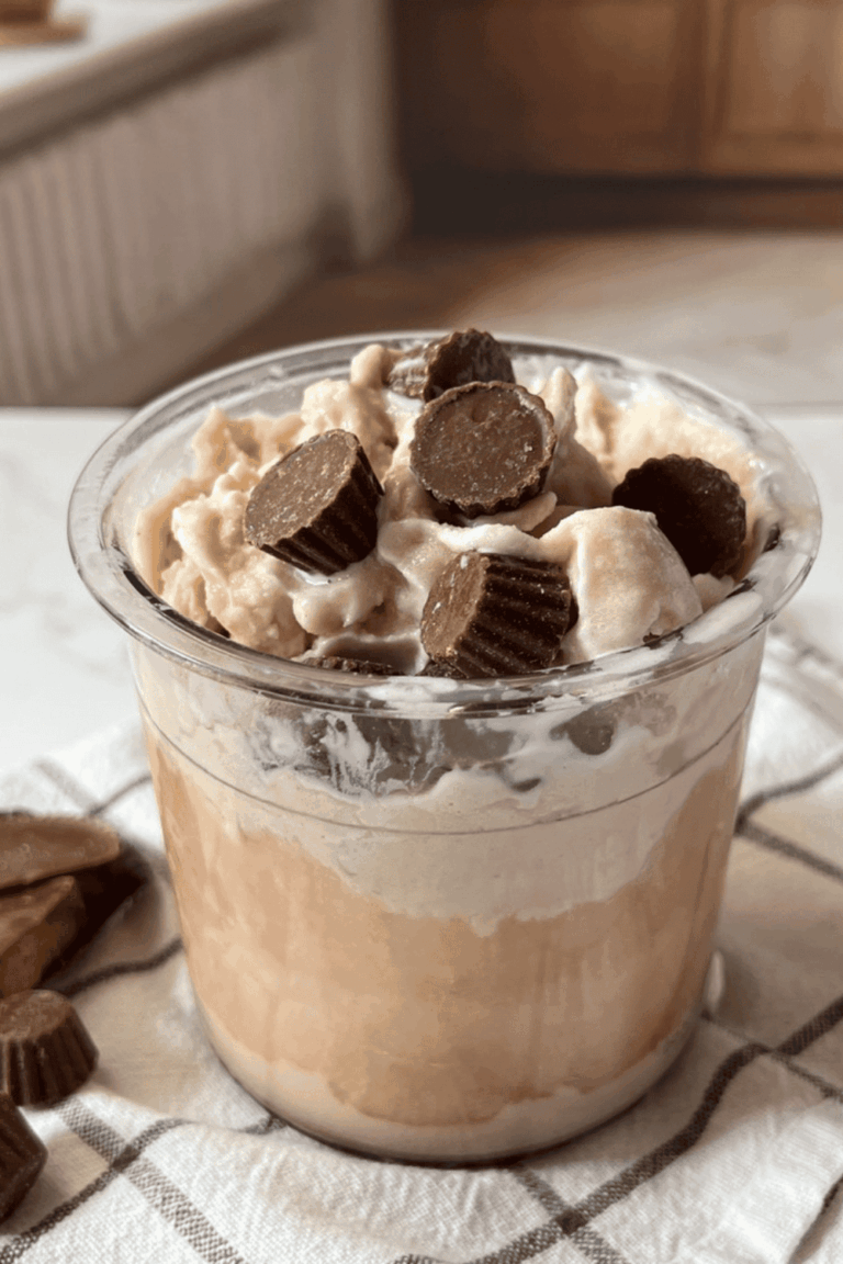 A clear Ninja Creami pint container overflowing with creamy chocolate peanut butter protein ice cream, topped with mini Reese's peanut butter cups, sitting on a white and gray plaid kitchen towel with extra peanut butter cups scattered alongside on a white surface.