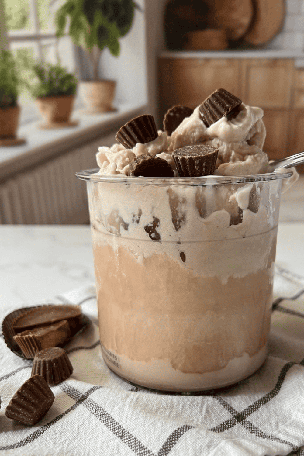 A clear Ninja Creami pint filled with layers of chocolate peanut butter protein ice cream and topped with mini Reese's peanut butter cups, sitting on a white and gray plaid kitchen towel with extra peanut butter cups scattered alongside and potted green plants on a sunny windowsill in the background.
