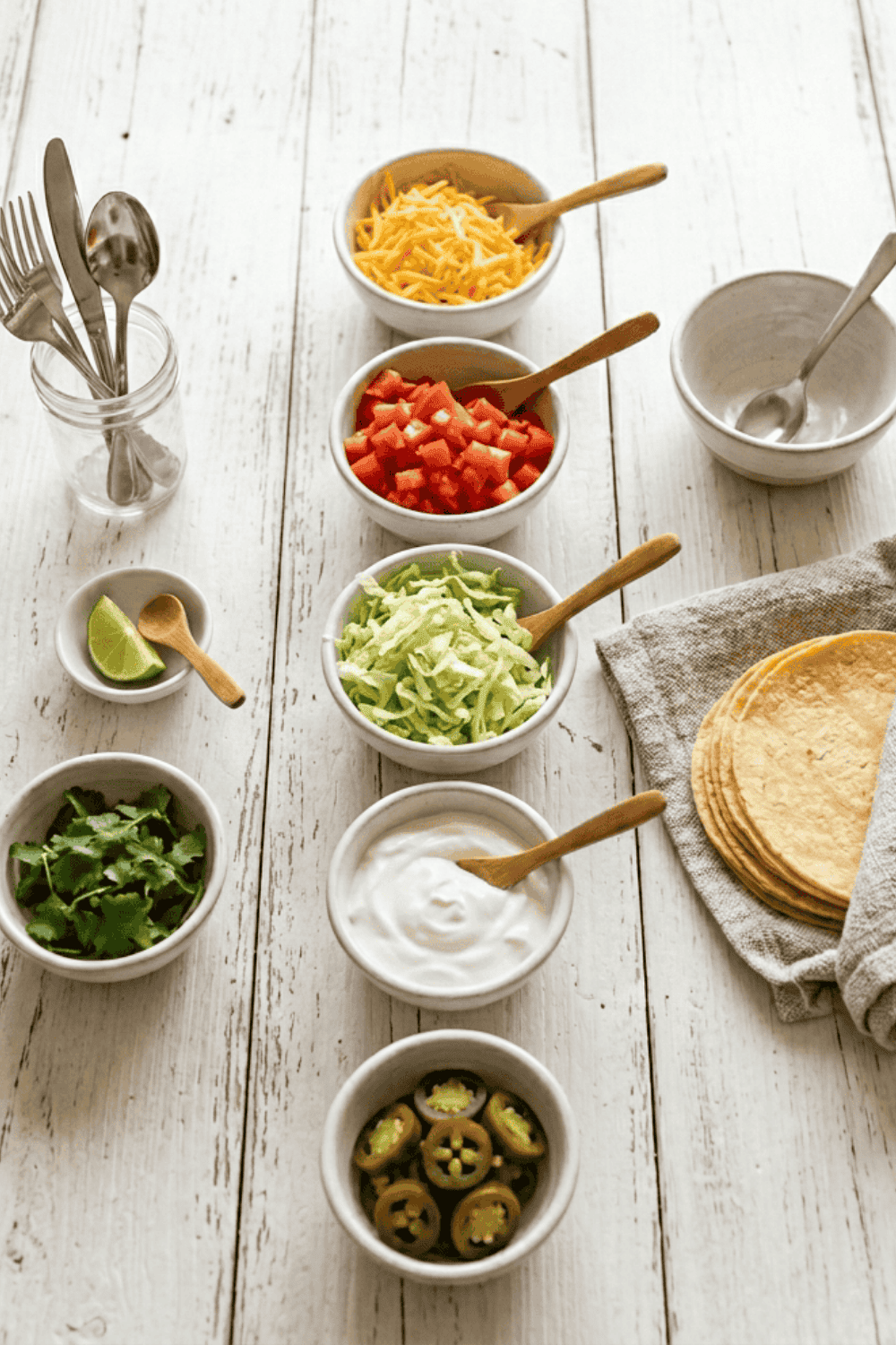 Overhead view of taco bar set up ideas on a white wood surface with small white bowls of shredded cheddar cheese, diced tomatoes, shredded lettuce, sour cream, fresh cilantro, and sliced jalapeños, each with a small wooden spoon. A lime wedge, silverware in a glass jar, and a stack of corn tortillas on a gray linen napkin complete the taco bar buffet layout.