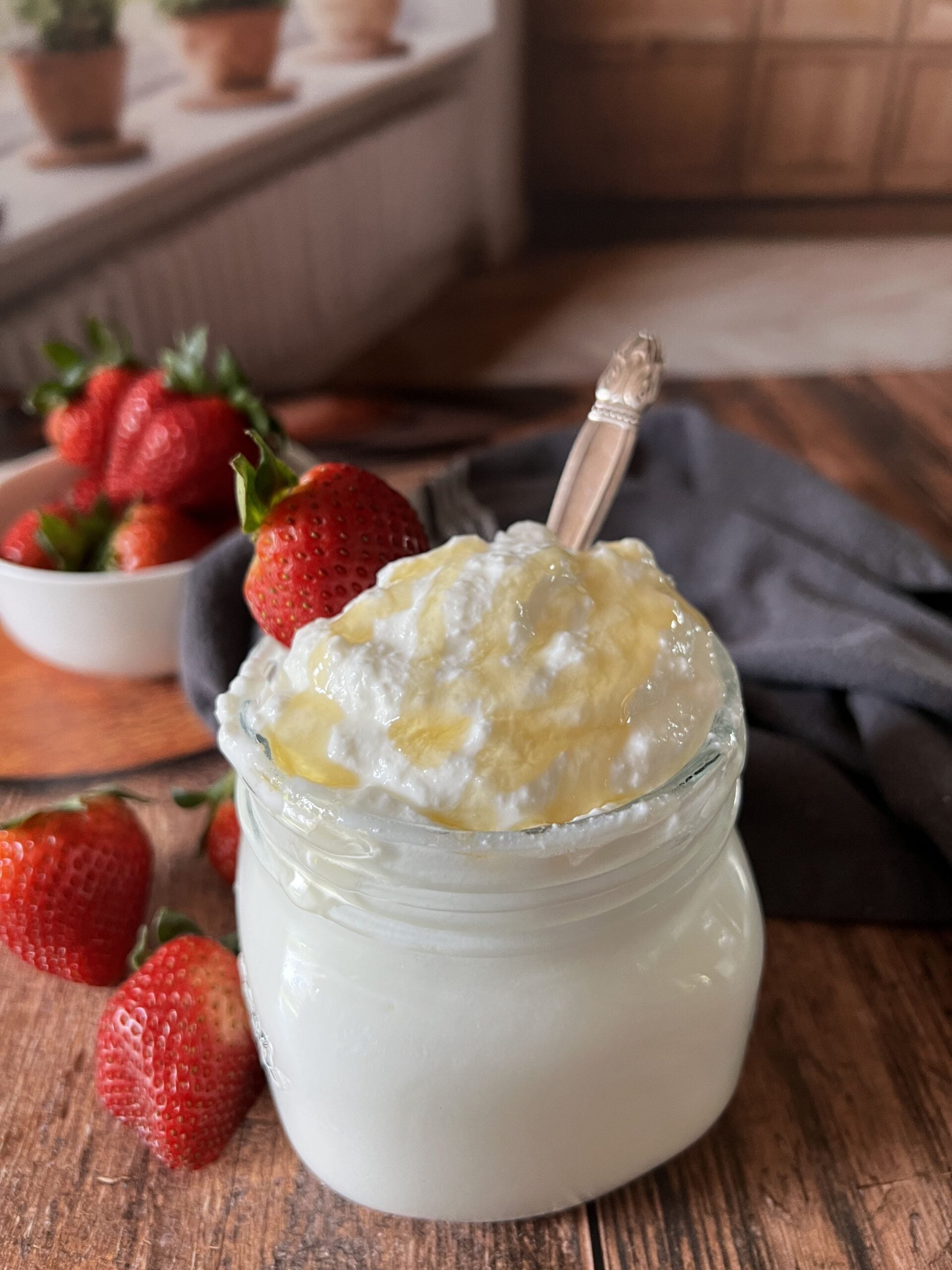 A glass jar of homemade Instant Pot Greek yogurt topped with honey and a vintage silver spoon, surrounded by fresh strawberries on a warm wood surface with terracotta herb pots on a sunny windowsill in the background