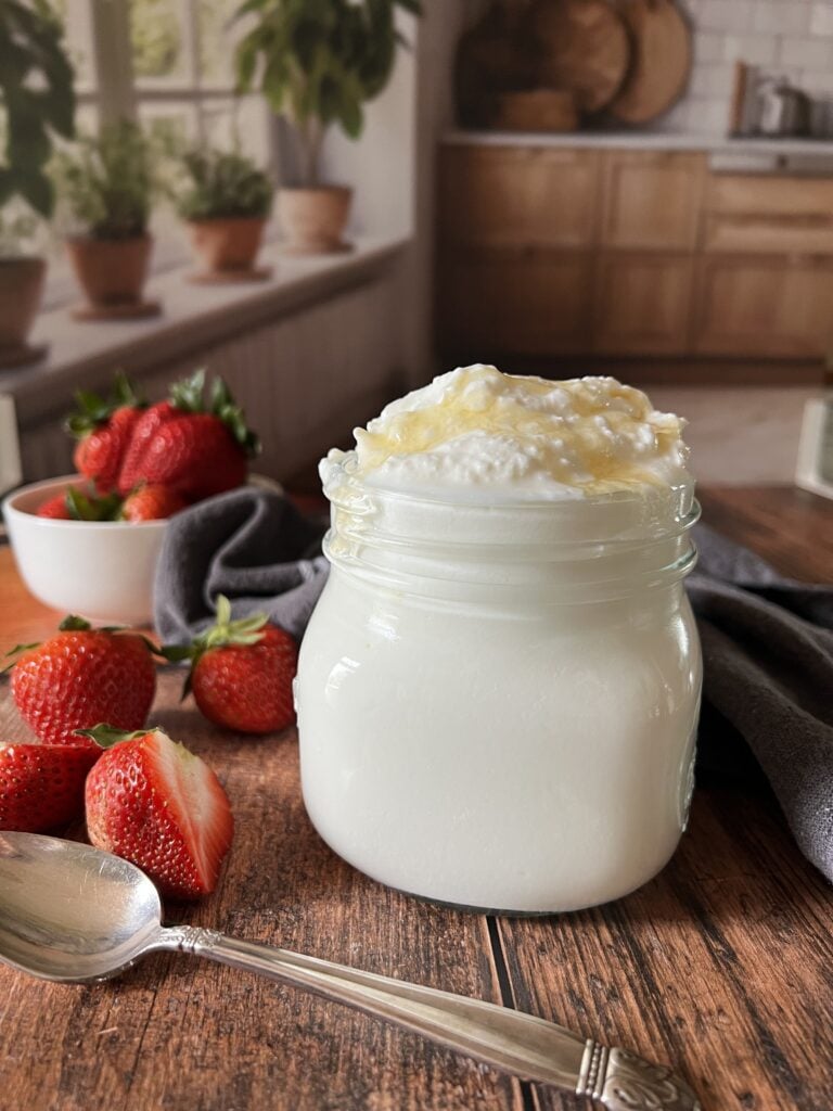 A glass jar of thick Instant Pot Greek yogurt topped with golden honey drizzle, surrounded by fresh red strawberries and a vintage silver spoon on a dark rustic wood surface with herb pots and a warm kitchen.