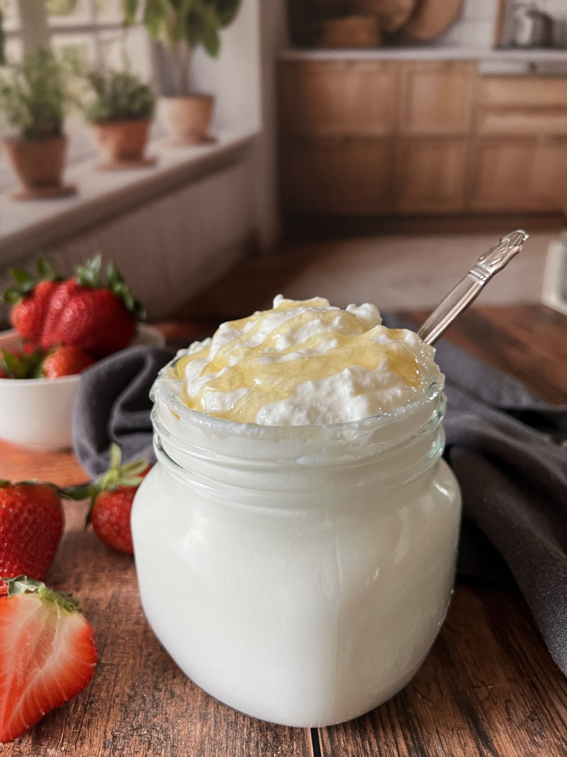 A glass jar of creamy homemade Greek yogurt with golden honey and a vintage silver spoon resting inside, surrounded by fresh strawberries on a rustic wood board with a white bowl of berries and a soft kitchen backdrop, easy Instant Pot yogurt recipe