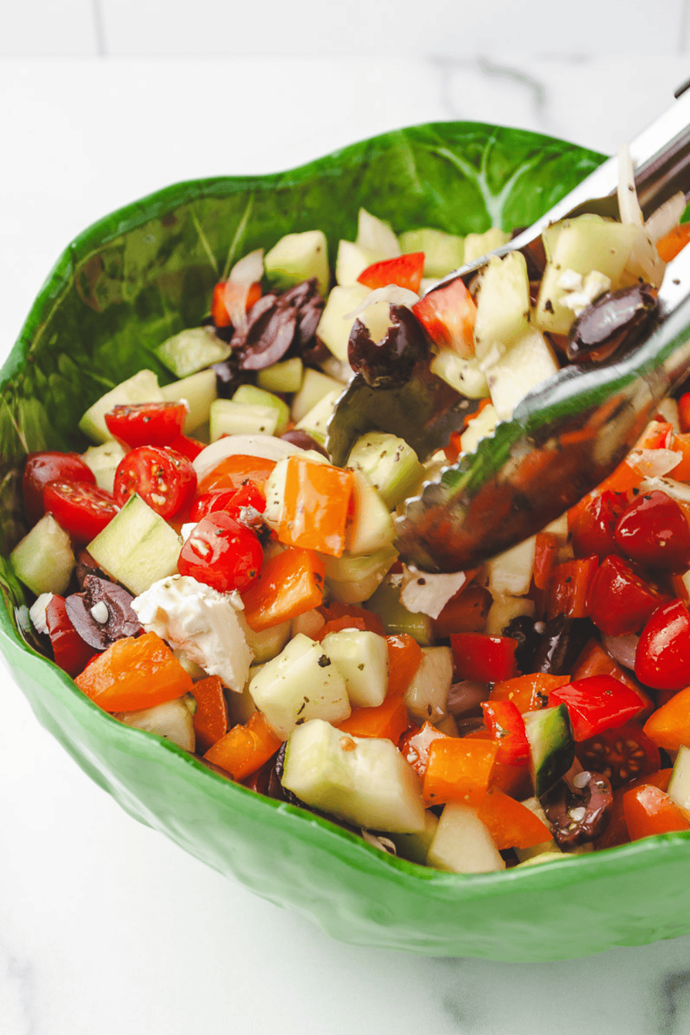 Classic Greek salad recipe being tossed with silver tongs in a green leaf-shaped bowl on a white marble surface, showing diced cucumbers, grape tomatoes, red and orange bell peppers, Kalamata olives and crumbled feta cheese with homemade Greek salad dressing.