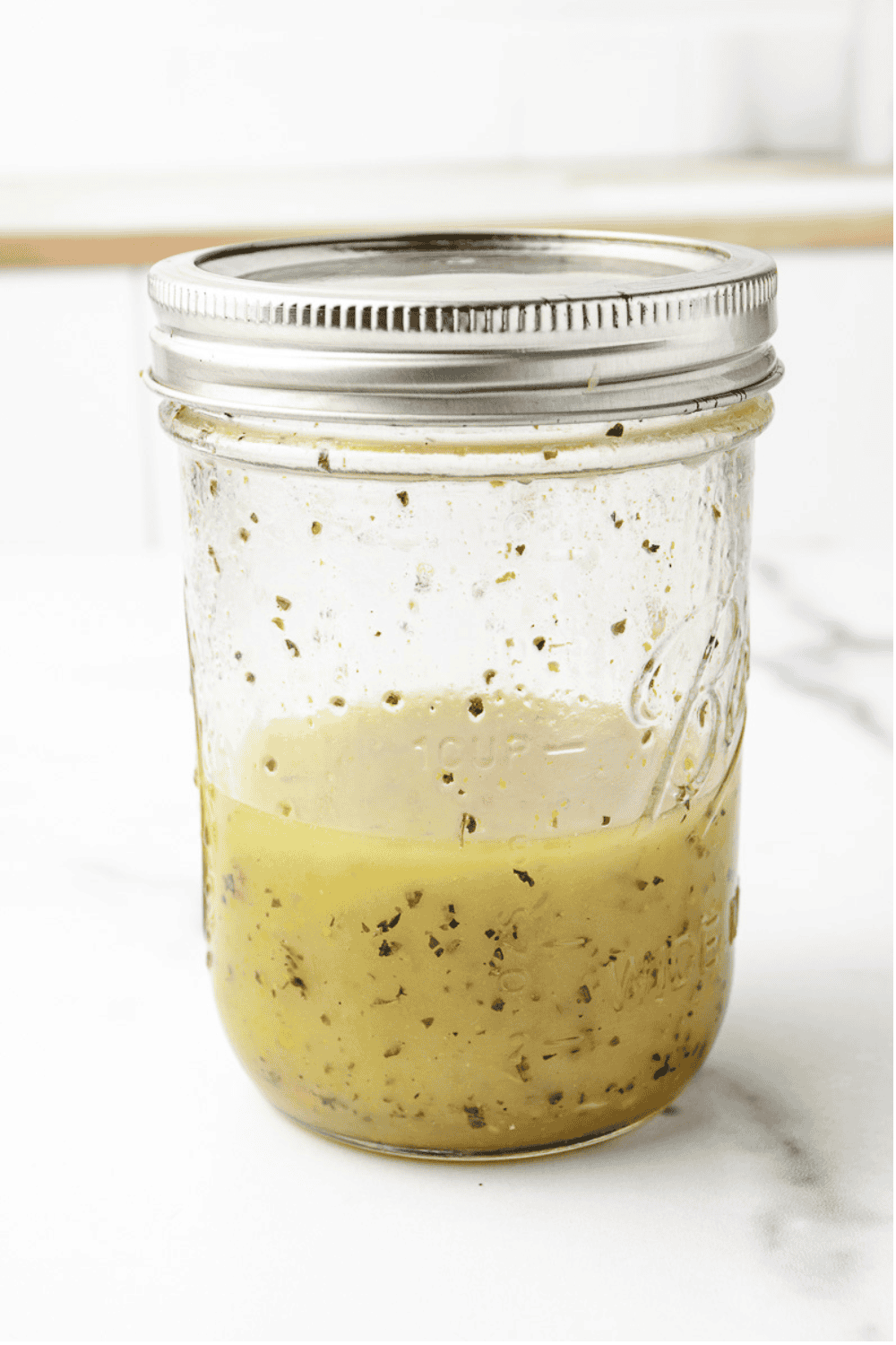 Homemade Greek salad dressing with herbs and oregano in a glass mason jar with a silver lid on a white marble surface.